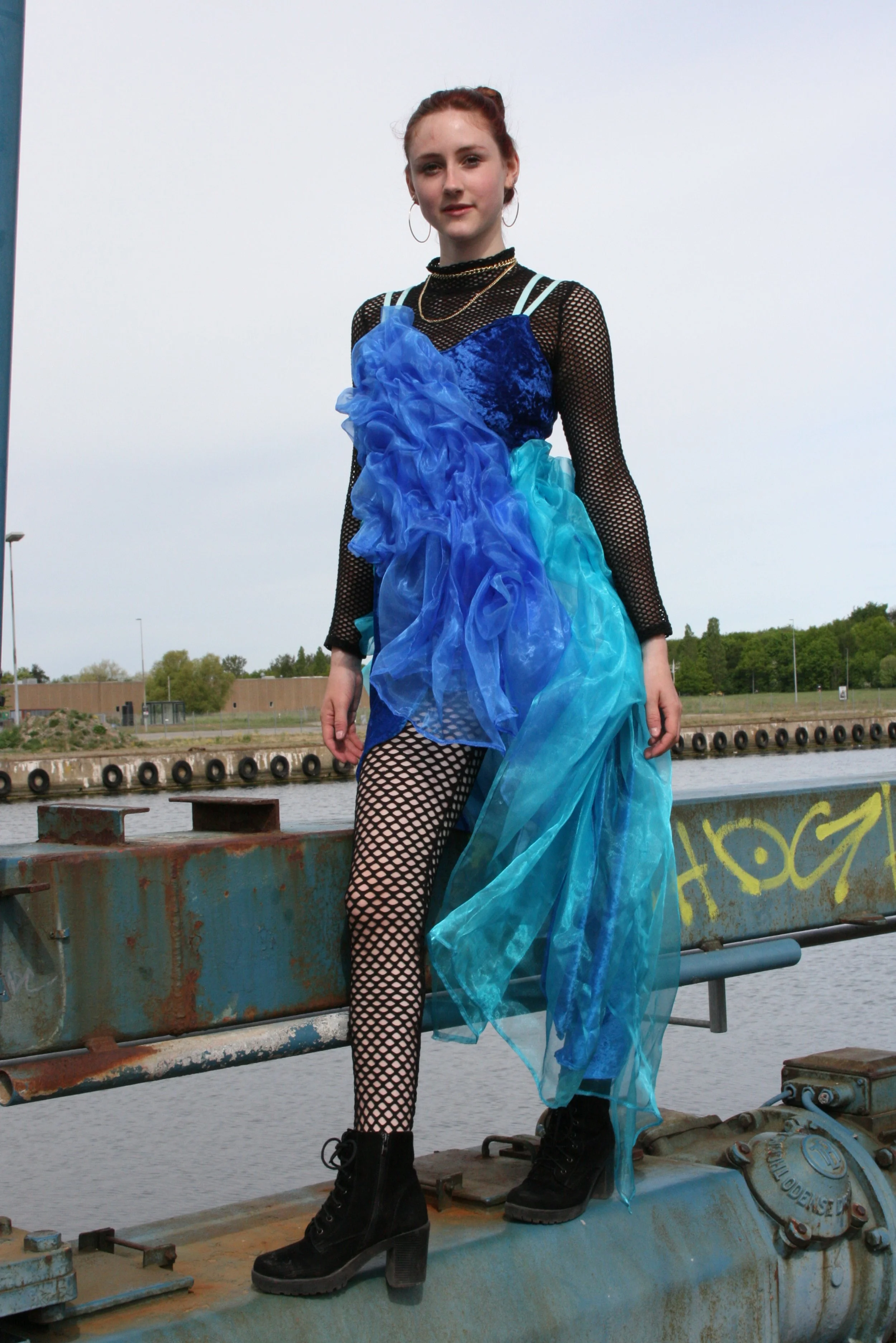 Young woman standing on a rusted industrial platform by water, wearing a layered blue dress with fishnet sleeves, black fishnet tights, and black lace-up boots.