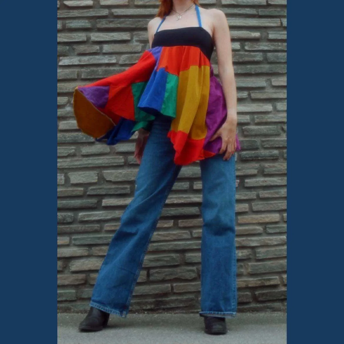 Person wearing a black strapless top, jeans, and black shoes, holding a large, colorful, rainbow-patterned fabric garment in front of a brick wall.
