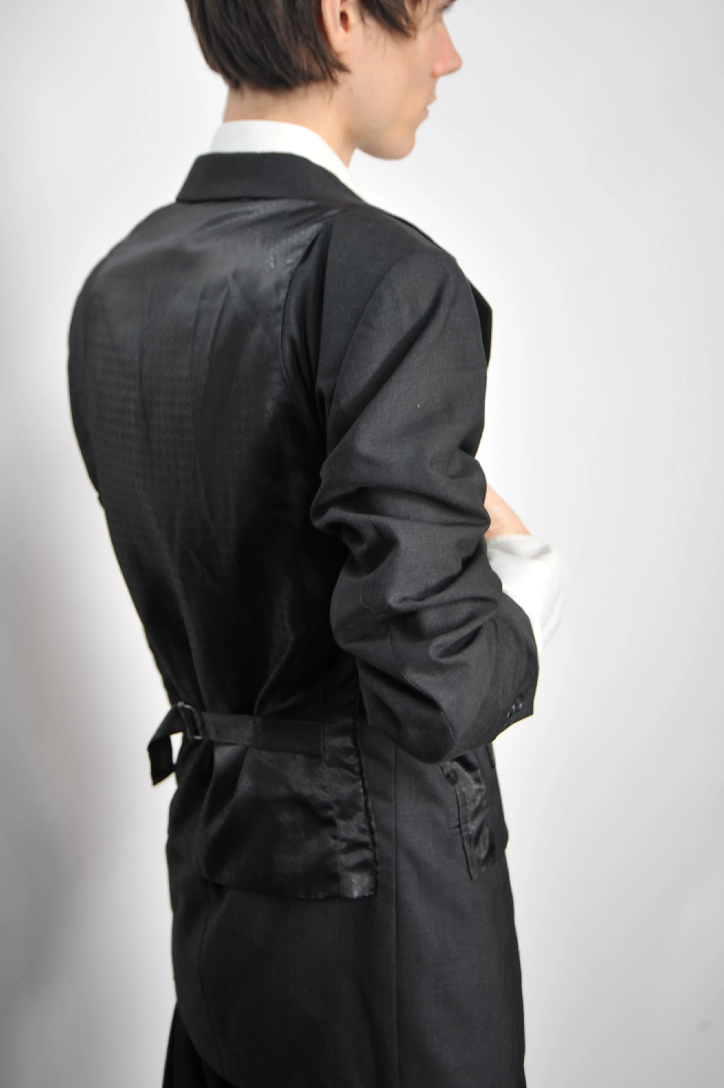 A person wearing a black formal jacket with a belt, turned sideways with arms crossed, against a plain white background.