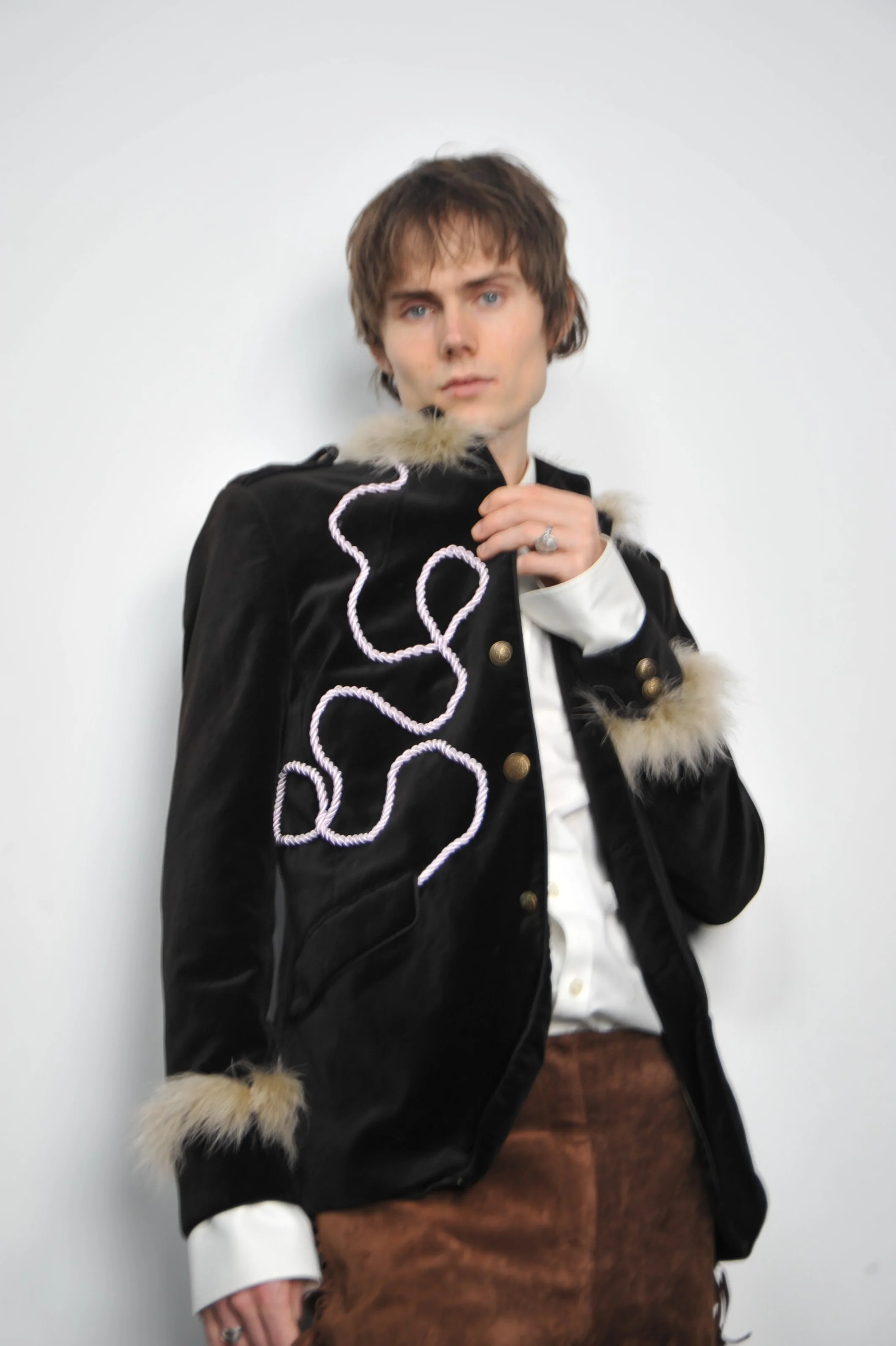 A young man with brown hair and blue eyes, wearing a black jacket with fur trim and decorative white piping, standing against a plain white background.