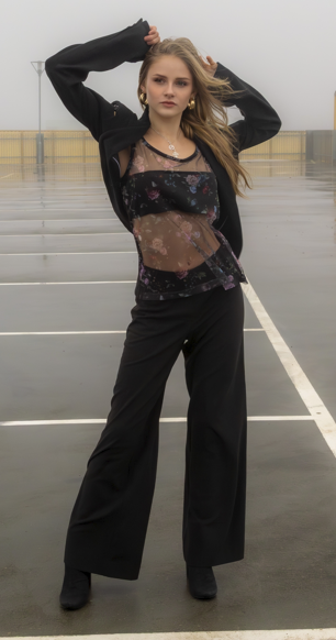 Young woman standing on an empty rooftop parking lot on a rainy day, holding her hair with both hands, wearing a black jacket, sheer decorated top, and wide-leg pants.