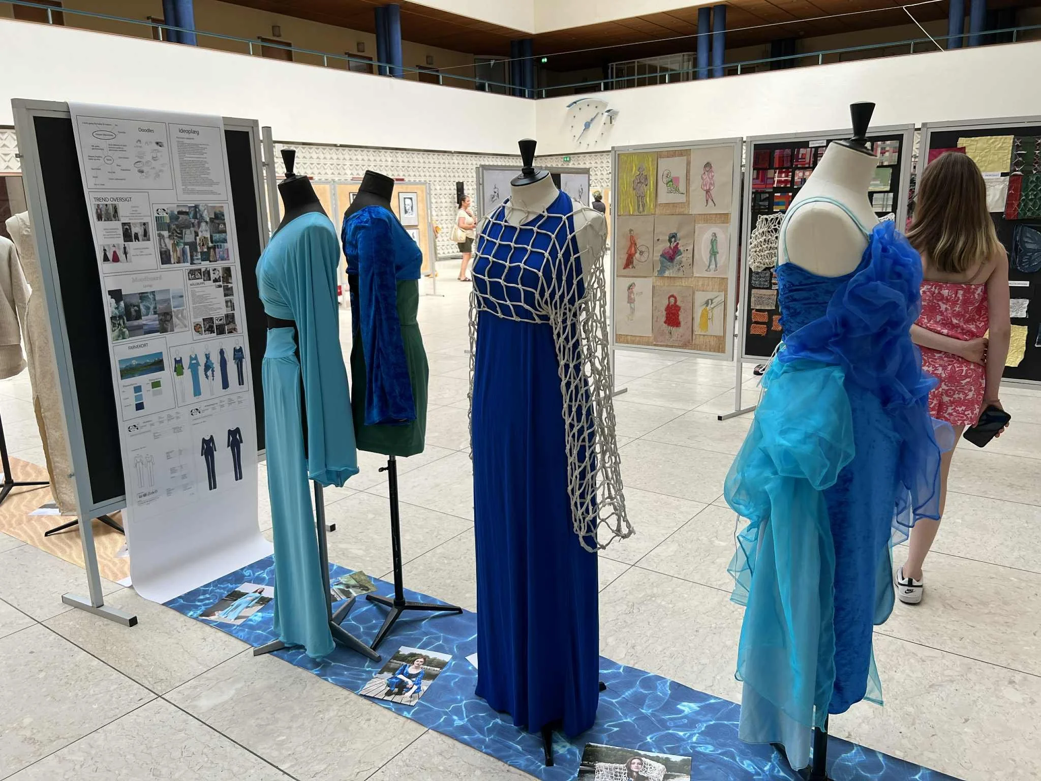 Display of mannequins dressed in blue and green dresses at an art and fashion exhibit, with informational boards and artwork in the background.