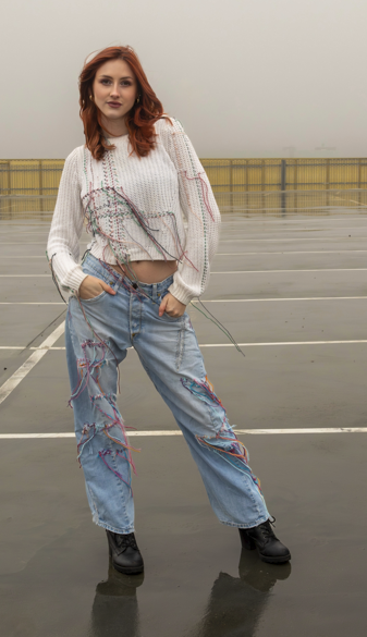 A young woman with red hair standing in an empty parking lot, wearing a white sweater and distressed jeans with colorful embroidery, and black boots.