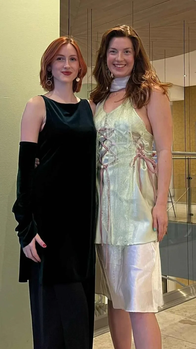 Two women standing together, smiling, in an indoor setting with a modern background. One woman has shoulder-length red hair and is wearing a black sleeveless dress with black gloves. The other woman has long, wavy brown hair and is wearing a light-colored, shiny dress with lace and ribbon details.
