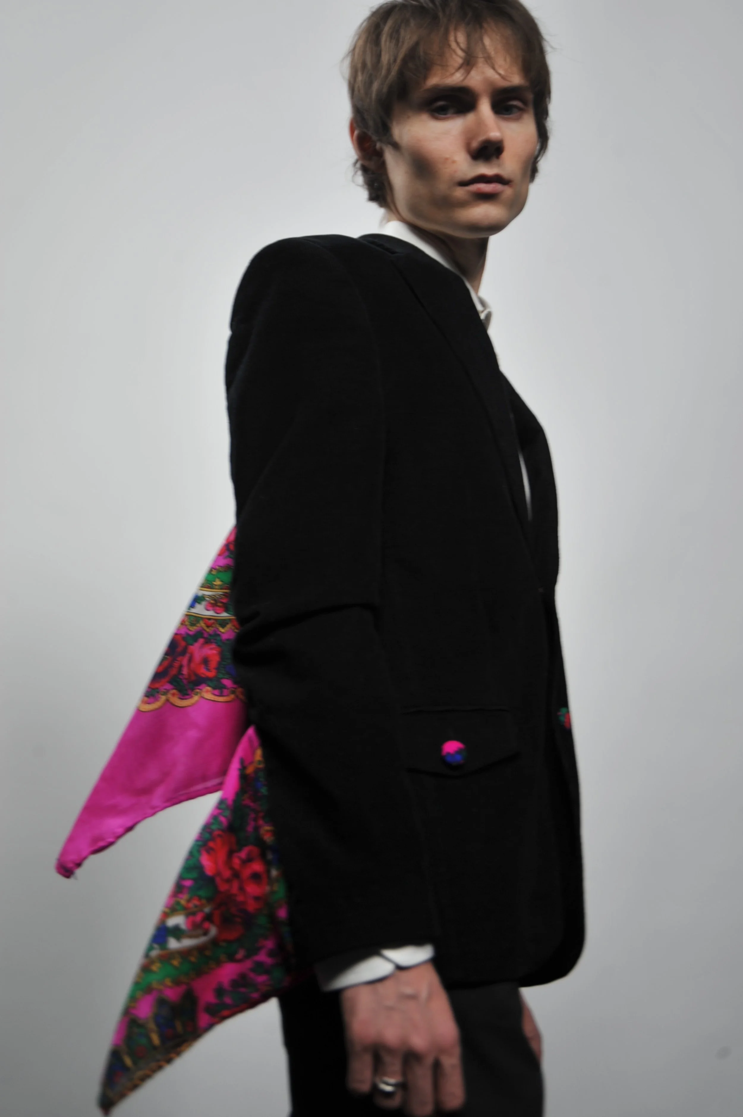 A young man with tousled hair wearing a black blazer and white shirt, holding a colorful embroidered scarf or cloth draped over his shoulder, standing against a plain light gray background.