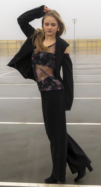 A young woman standing in an empty parking lot on a rainy day, wearing a black jacket, floral top, and black pants with her hand on her head.