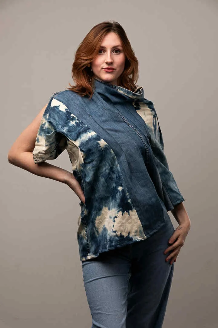 A woman with shoulder-length auburn hair in a blue and white tie-dye sleeveless top and blue jeans posing with one hand on her hip against a plain gray background.