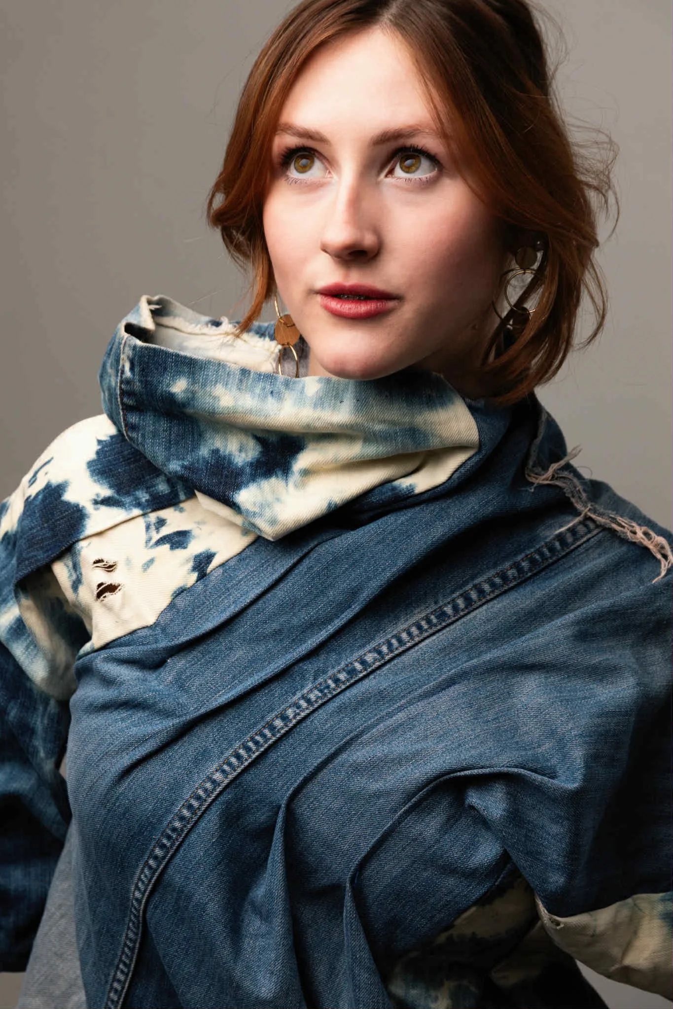 A young woman with short red hair, wearing a denim jacket and a tie-dye scarf, looking upward.