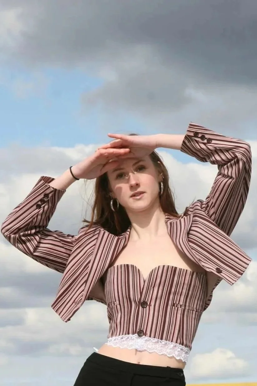 A young woman with long red hair holding her hand above her eyes, looking into the distance against a cloudy sky.
