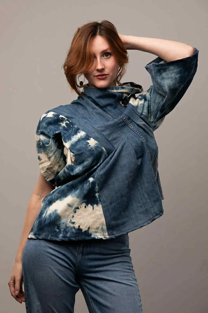 A woman with red hair posing with her hand behind her head, wearing a denim jacket with a tie-dye pattern and matching jeans against a plain gray background.