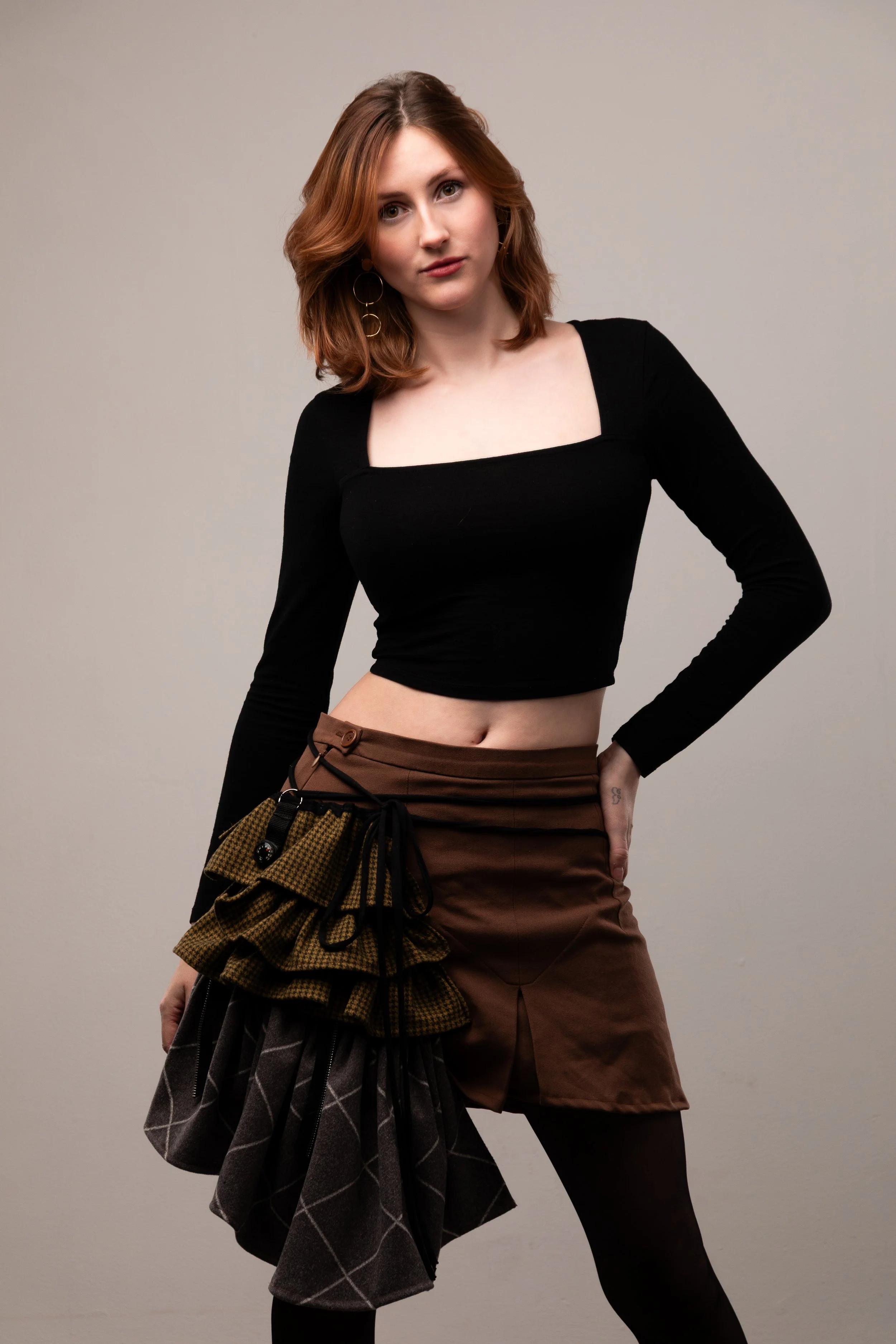 A young woman with red hair wearing a black long-sleeve crop top, brown skirt, and patterned fabric accessories, standing against a plain background.