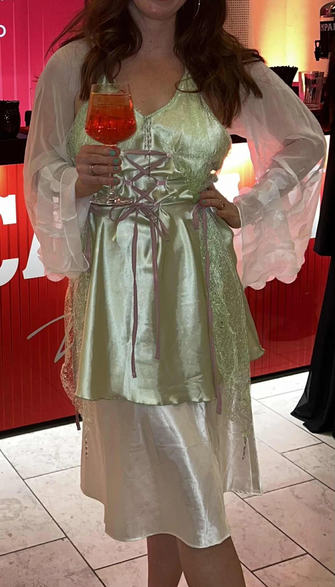 A woman holding a cocktail glass with an amber-colored drink, dressed in a shiny, light-colored dress with sheer, puffed sleeves, in an indoor setting with a red wall in the background.