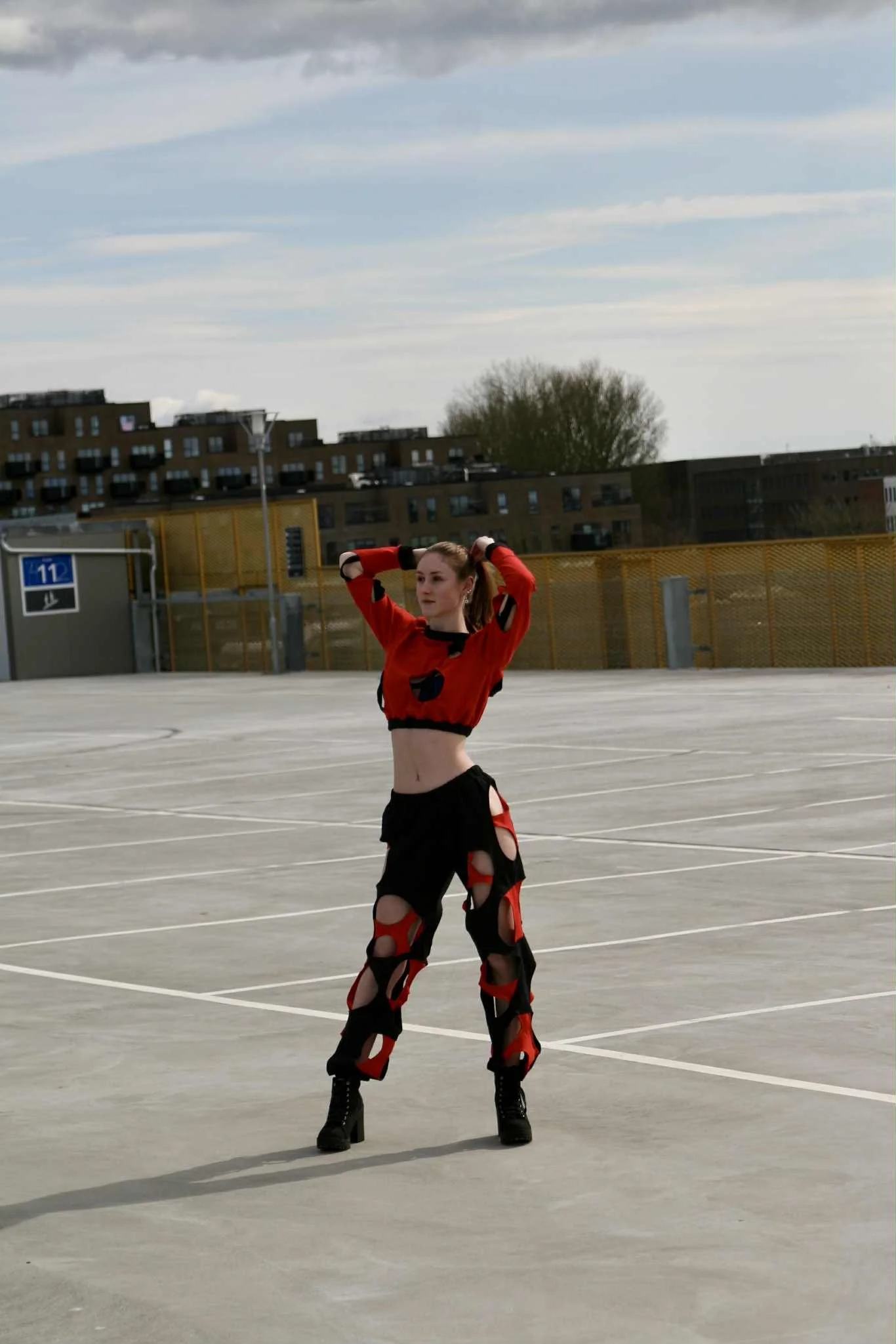 A woman in a red and black outfit with cutouts, wearing black platform boots, standing on an empty outdoor parking lot with her arms raised behind her head.