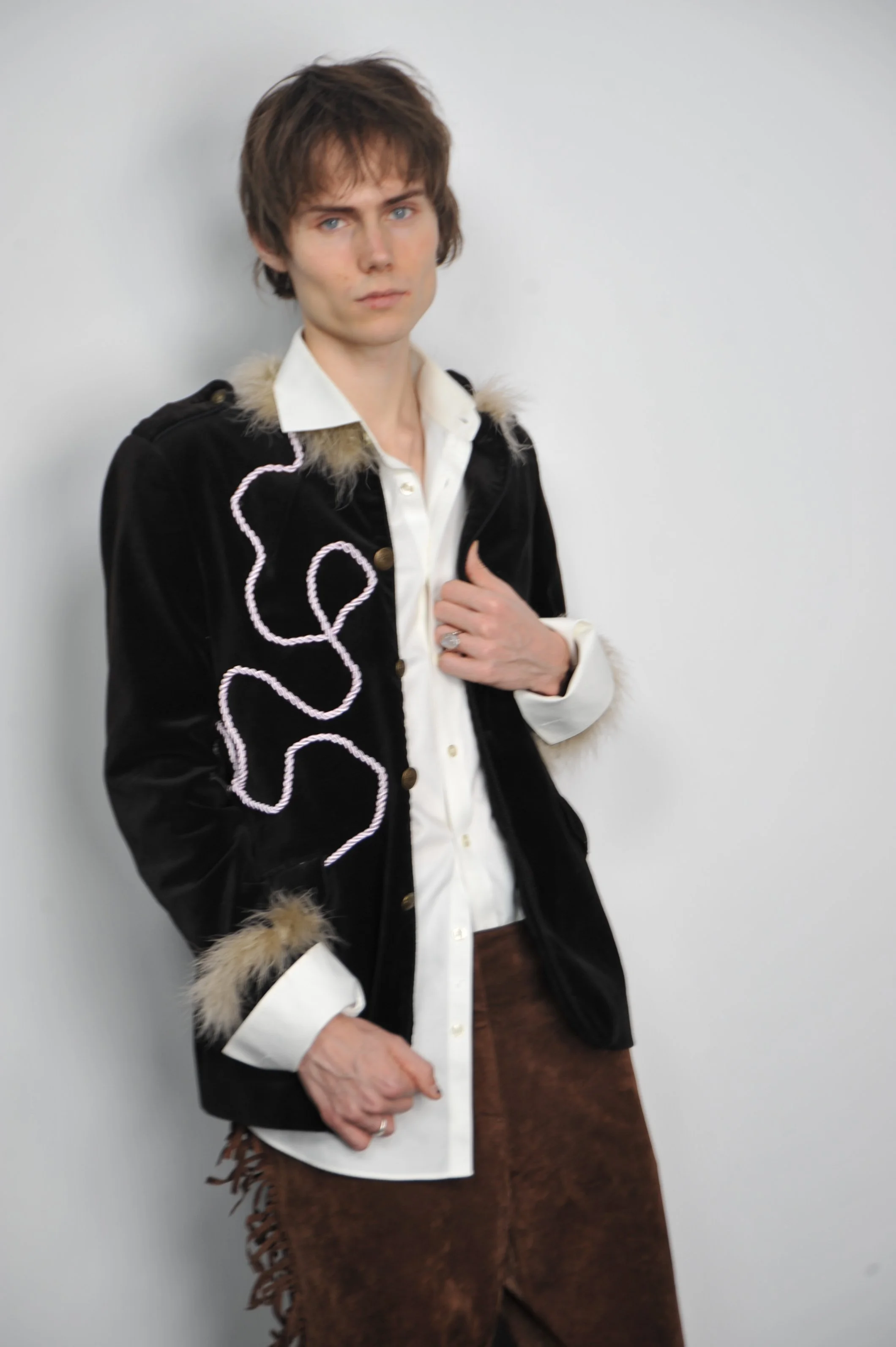 A young man with brown hair and blue eyes wearing a white shirt, black jacket with fur trim and decorative rope accents, and brown fringed pants. He is standing against a plain white wall, looking directly at the camera with a serious expression.
