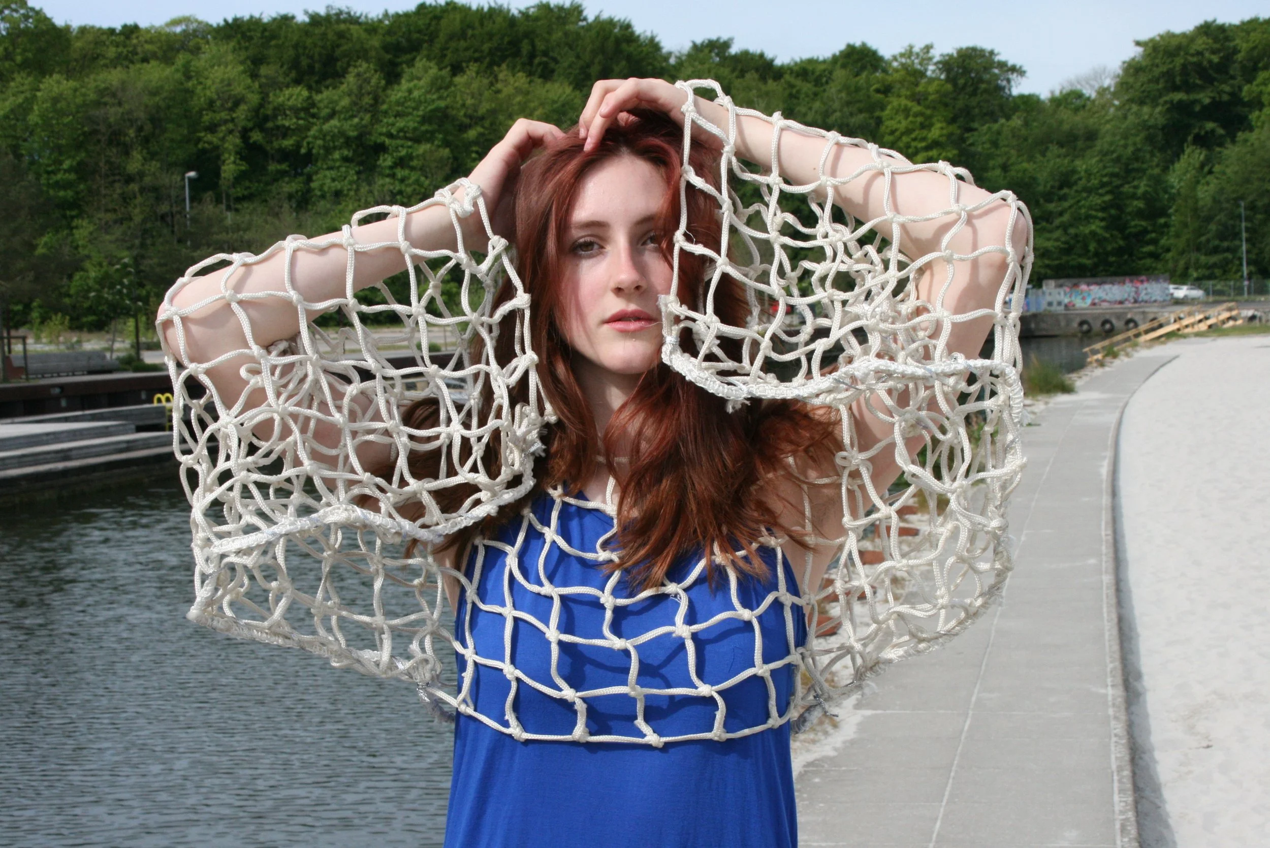 A young woman with red hair in a blue dress holding a large white net over her shoulders, standing on a riverside walkway with greenery in the background.
