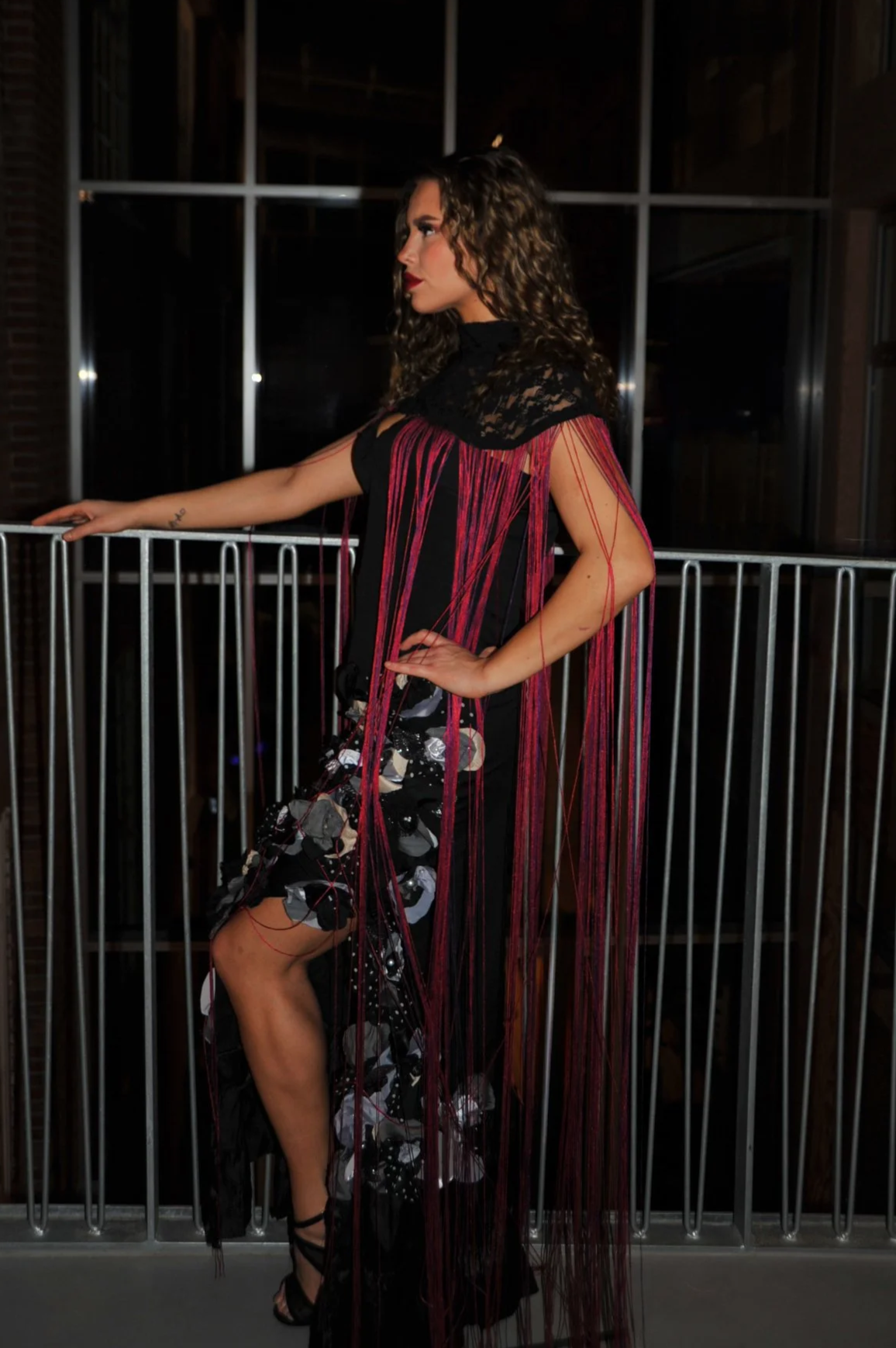 Woman with curly hair wearing a black dress with red strings and patterned fabric, standing on a balcony at night.