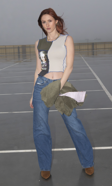 Young woman standing in an empty rooftop parking lot on a cloudy day, wearing a sleeveless graphic crop top, high-waisted jeans, and brown boots, holding a jacket.