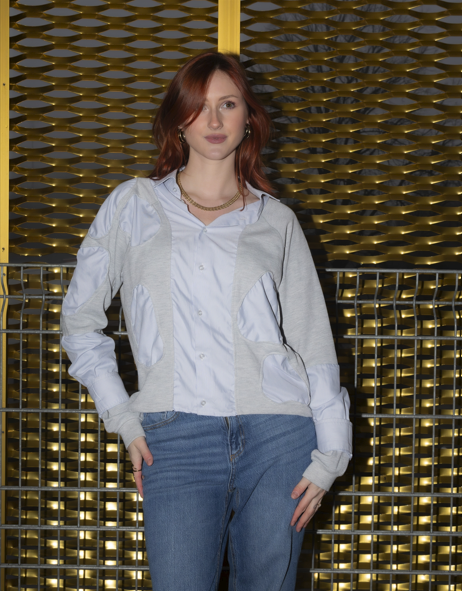 A young woman with red hair and gold jewelry standing in front of a gold decorative wall, wearing a white shirt, a light gray jacket, and blue jeans.