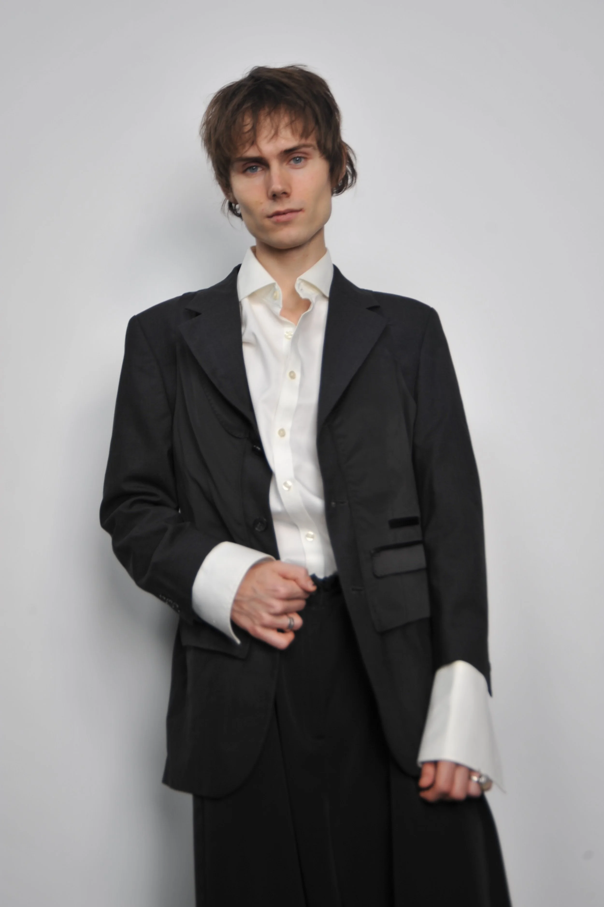 A young man with brown hair wearing a black suit and white shirt, standing against a plain white wall.