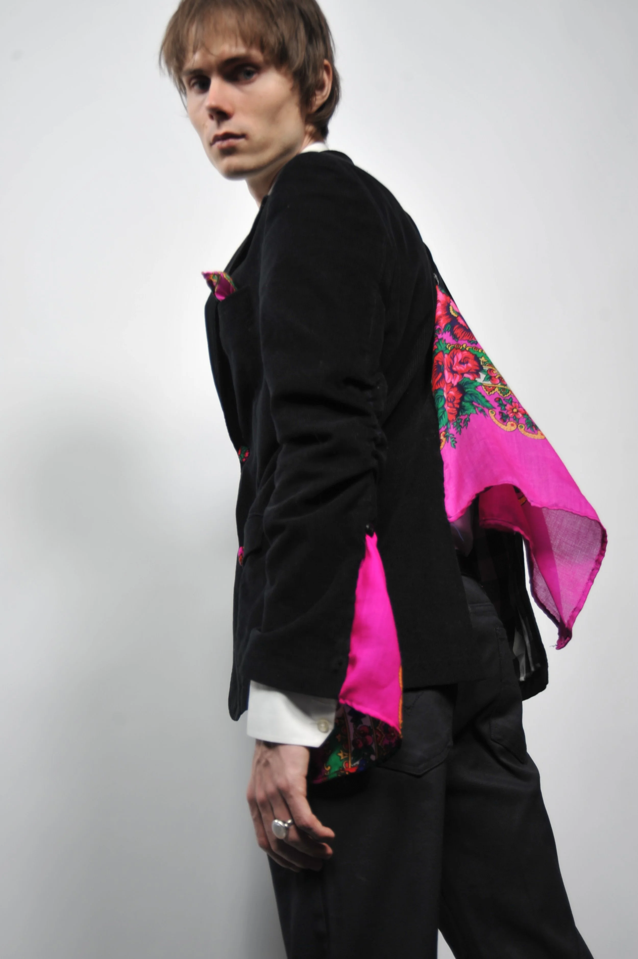 A young man with light brown hair wearing a black jacket with colorful floral accents and a bright pink scarf, standing against a plain white background.