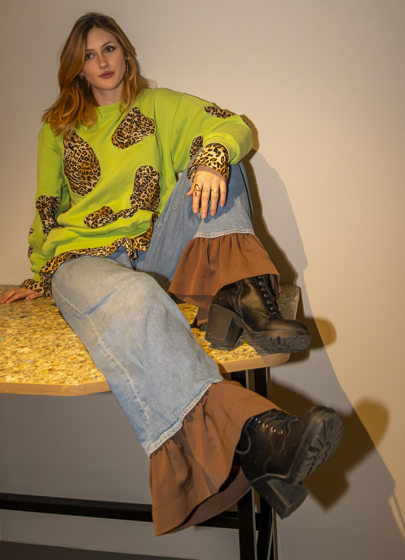 Young woman sitting on a table against a plain wall, wearing a neon green sweater with leopard print accents, light blue jeans, a layered brown skirt, and black combat boots.