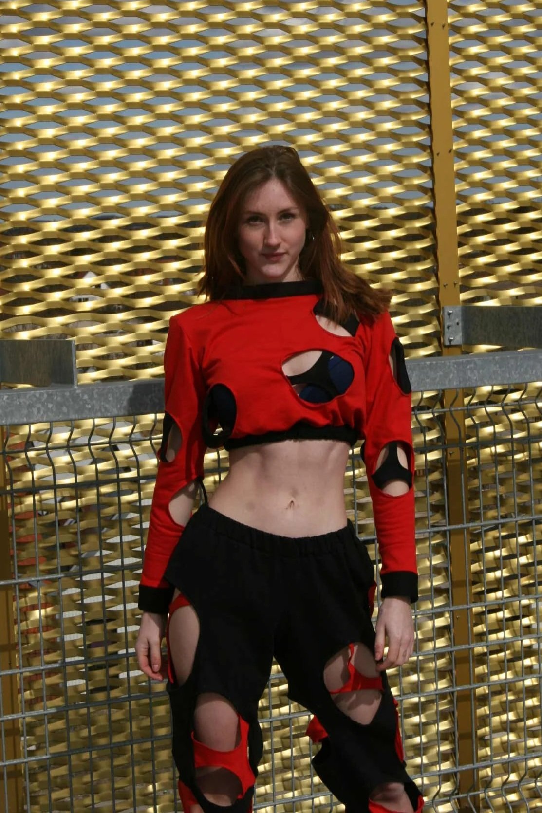 Young woman standing in front of a decorative golden wall, wearing a red and black ripped crop top and black ripped pants.