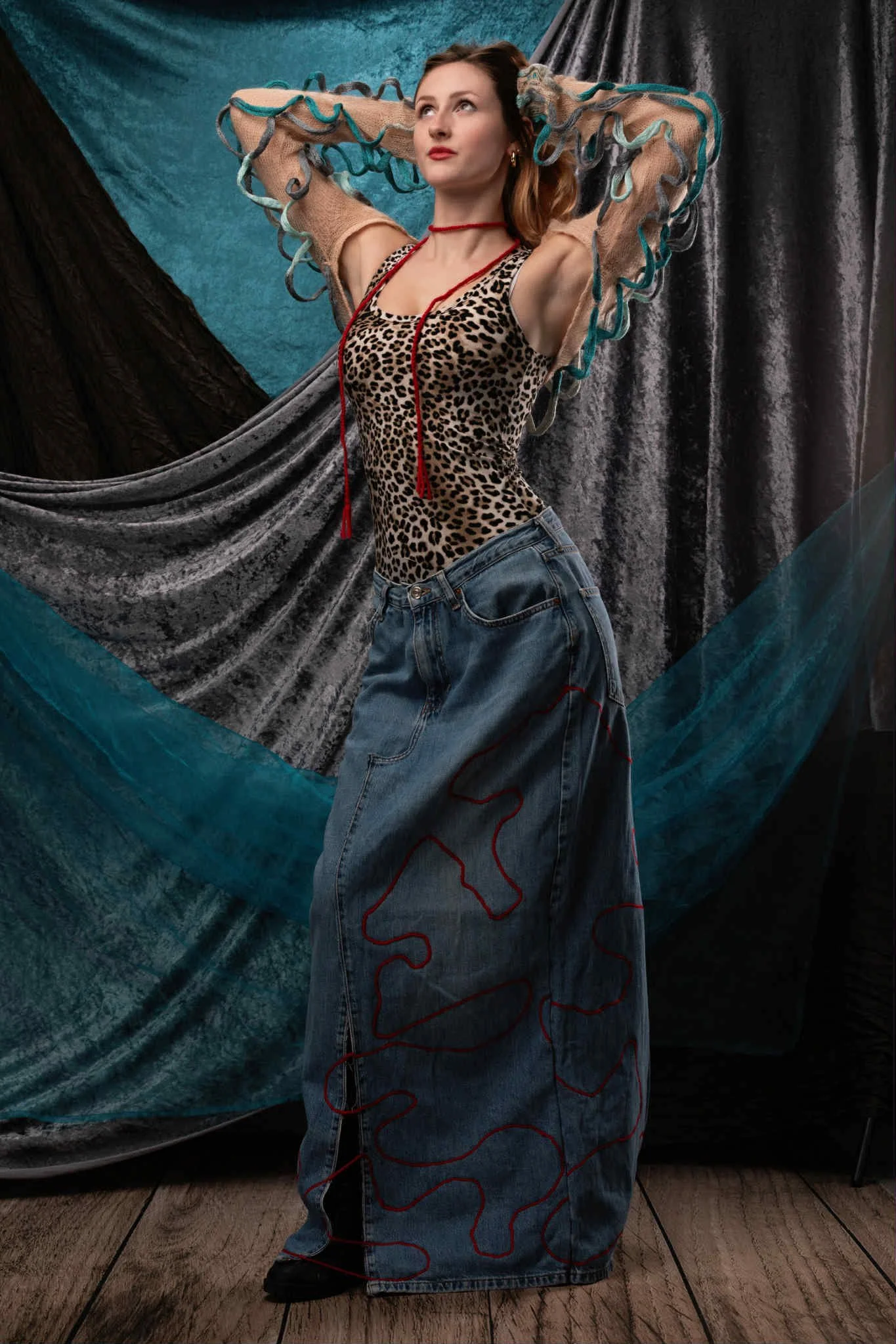 A woman with red hair wearing a leopard print top, jeans with embroidery, and transparent mesh sleeves stands with arms raised against a colorful fabric backdrop.
