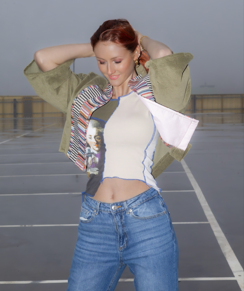 Young woman with red hair standing in an empty rooftop parking lot during daytime, adjusting her hair and wearing a colorful patchwork jacket over a graphic T-shirt and blue jeans.