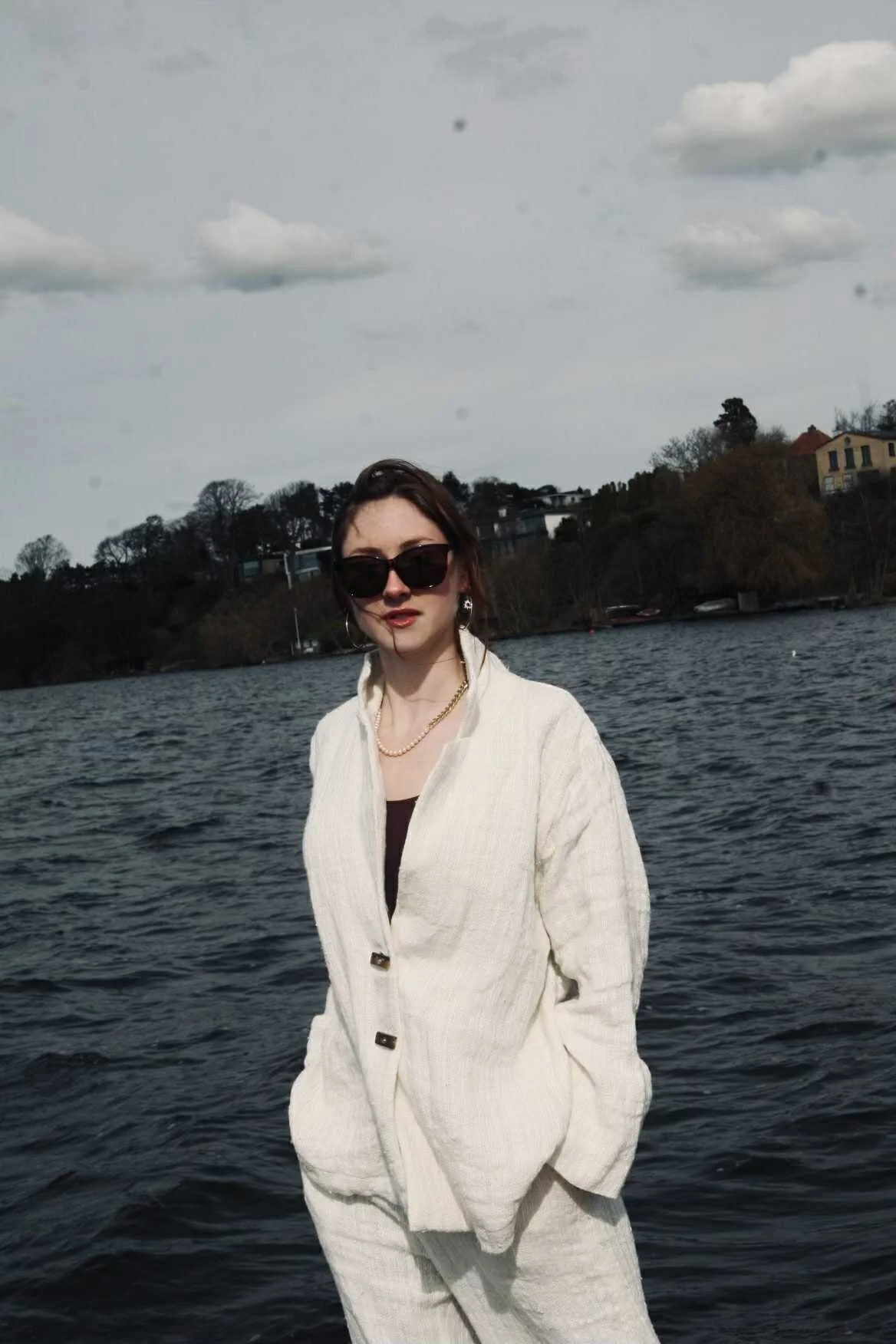A woman wearing a white suit and black shirt standing by a body of water, with trees and houses in the background under a cloudy sky.