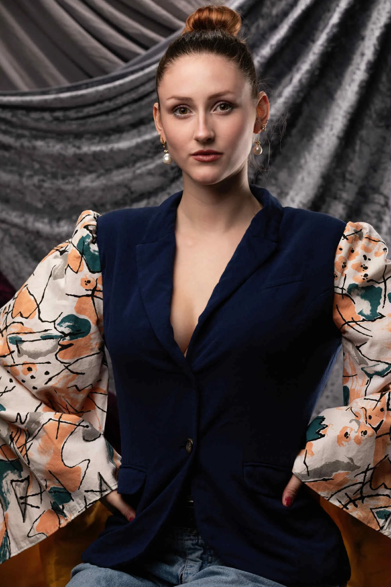 A woman with a high bun hairstyle, wearing pearl earrings, a navy blazer with floral patterned sleeves, and light blue jeans, standing against a textured gray and black backdrop.