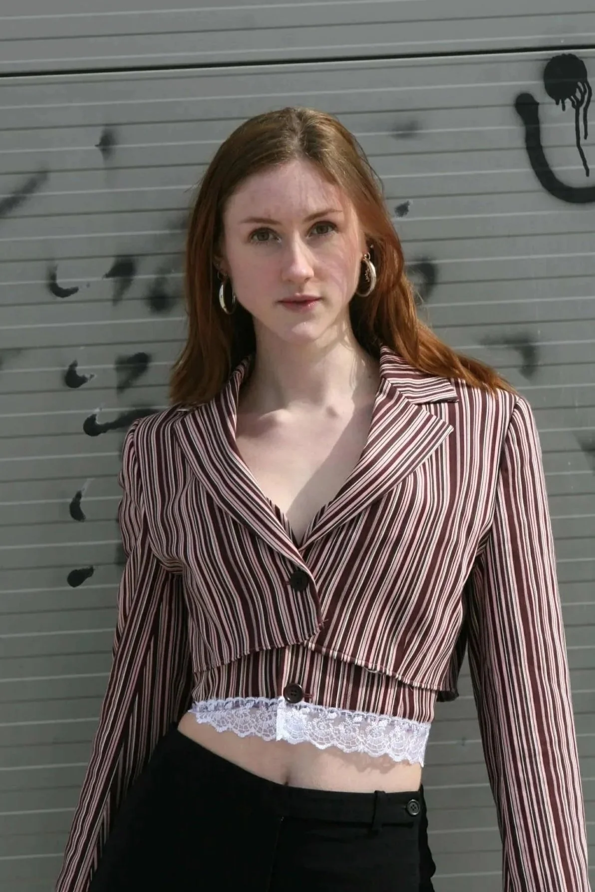 A young woman with red hair and hoop earrings standing in front of a gray, graffiti-covered wall, wearing a striped cropped blazer and white lace top.