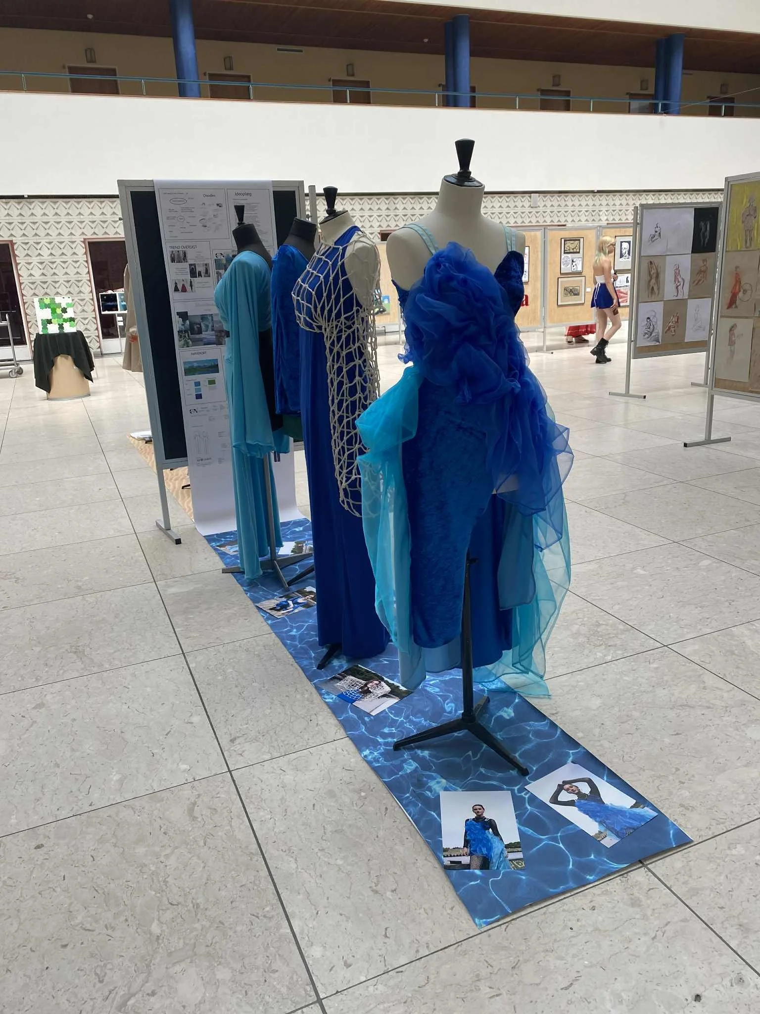 Display of blue fashion dresses and outfits on mannequins at an indoor exhibition, with fashion sketches and photographs on display panels and a blue water-patterned floor covering.