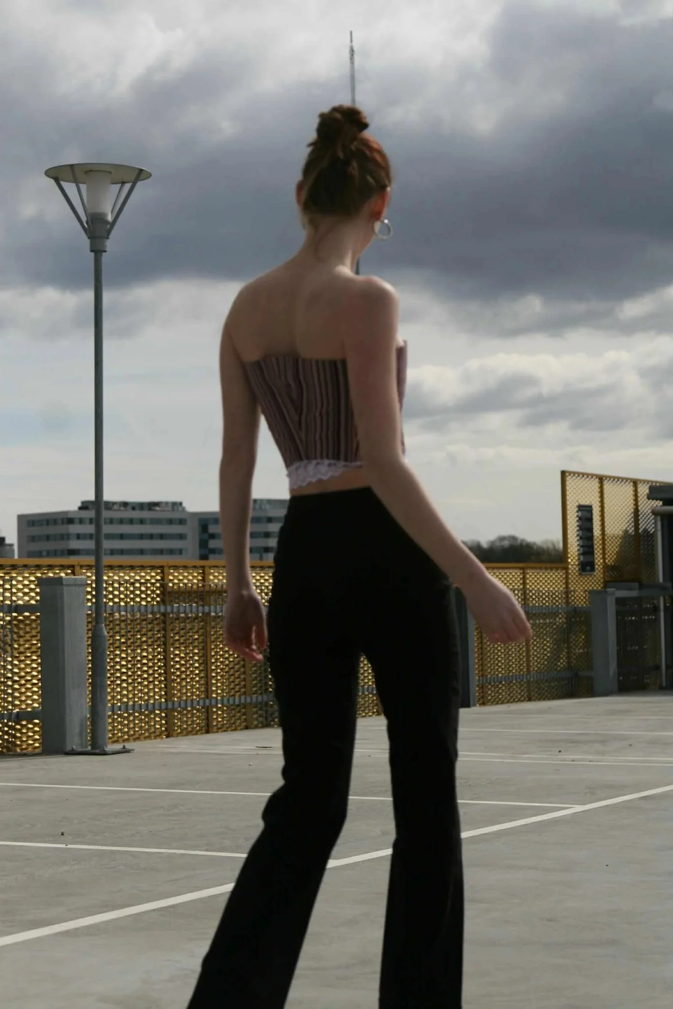 Woman with a high bun hairstyle, wearing a strapless striped top and black pants, walking on a parking garage rooftop under cloudy sky.