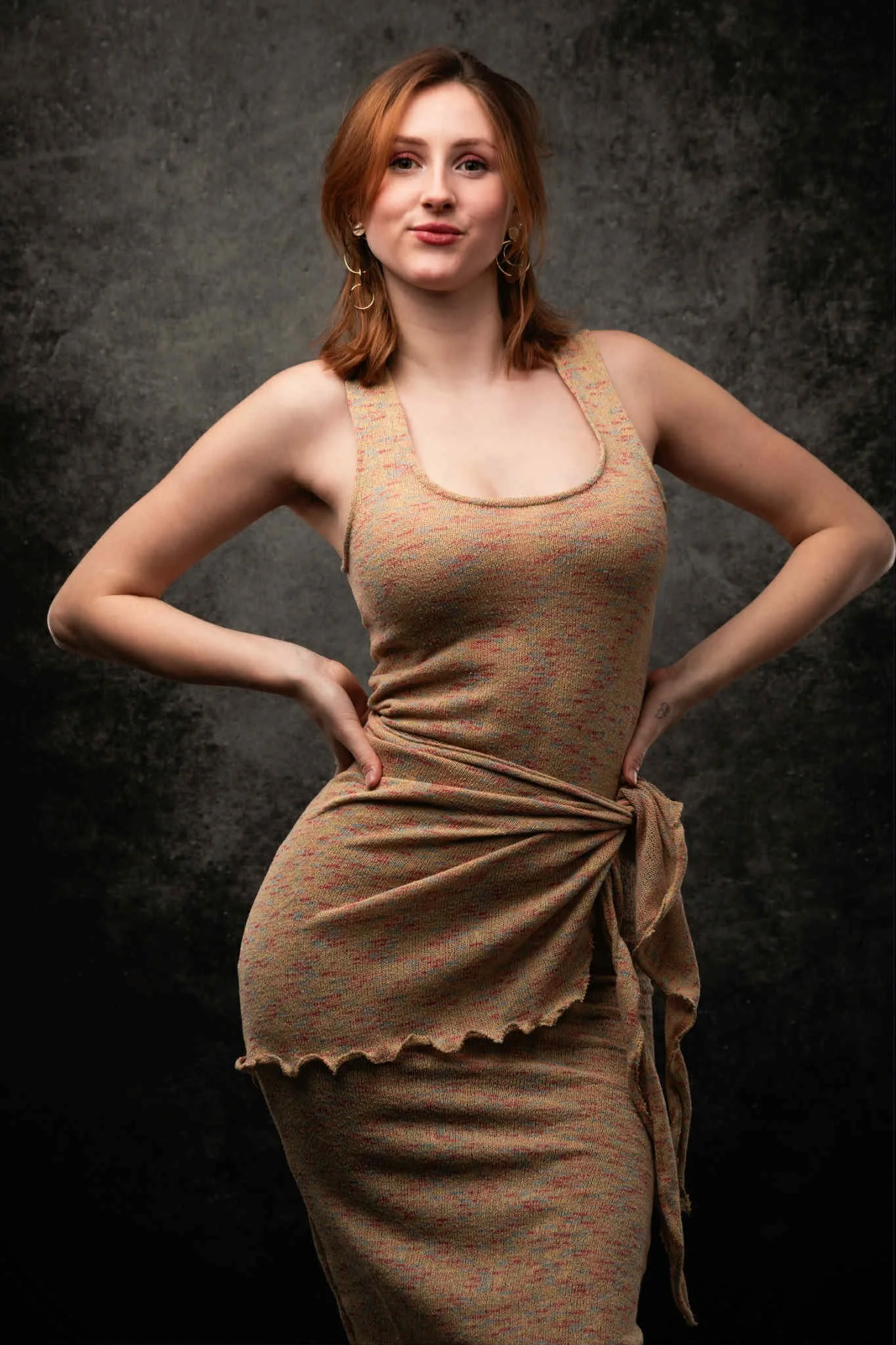 A woman with red hair standing with her hands on her hips overhead against a dark background.