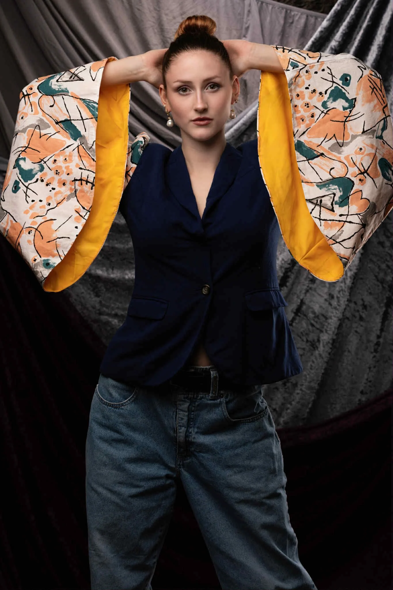 A young woman with red hair tied in a bun, wearing a navy blazer and gray jeans, posing with arms raised and hands behind her head, standing in front of a dark textured backdrop.