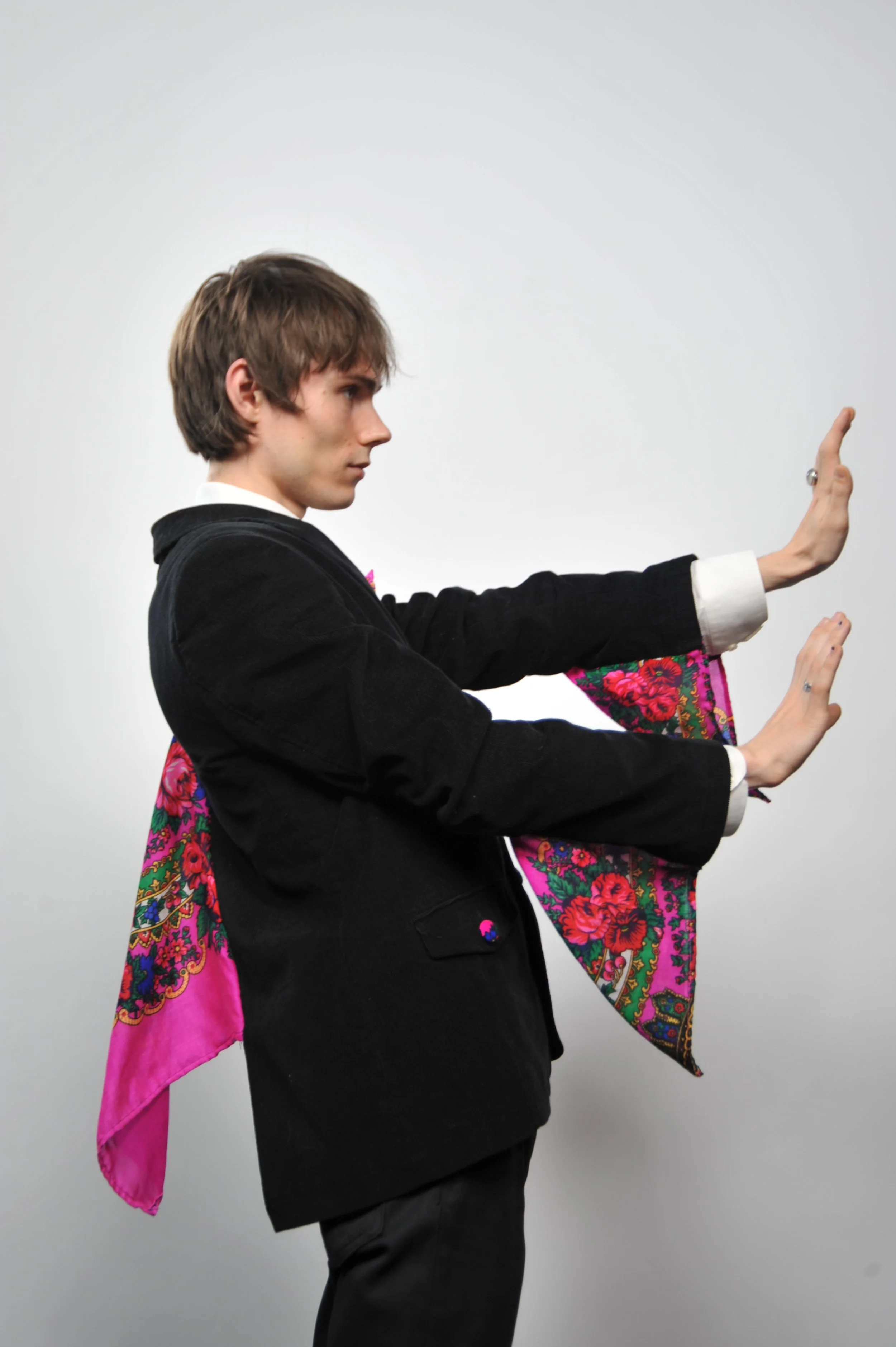 A young man in a black blazer and white shirt performs a Tai Chi pose against a plain white background, with one arm extended forward and the other bent, forearm perpendicular, with hands open and facing outward.