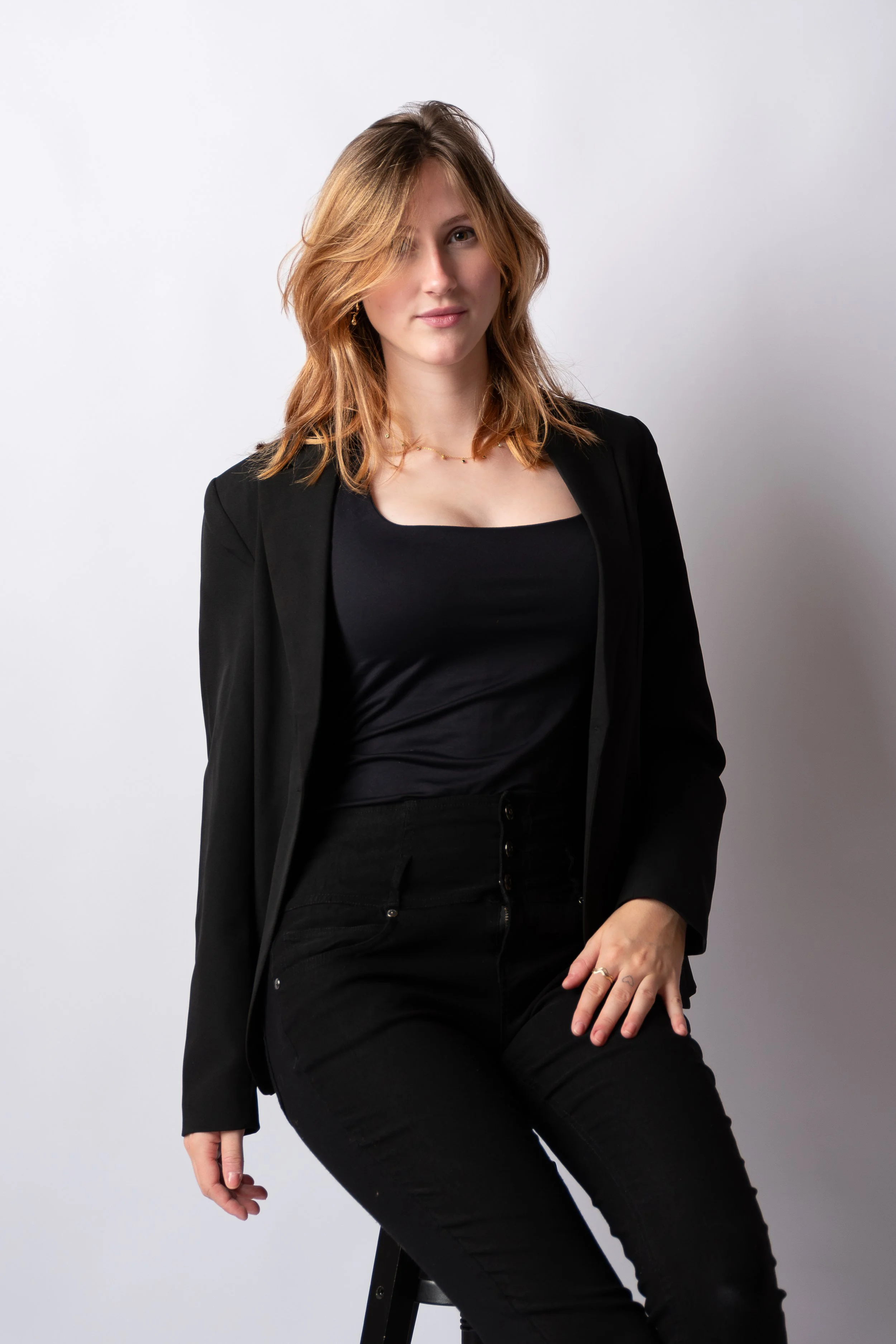 A woman with shoulder-length wavy red hair, wearing a black blazer, black top, and black pants, sitting on a stool against a plain light gray background.