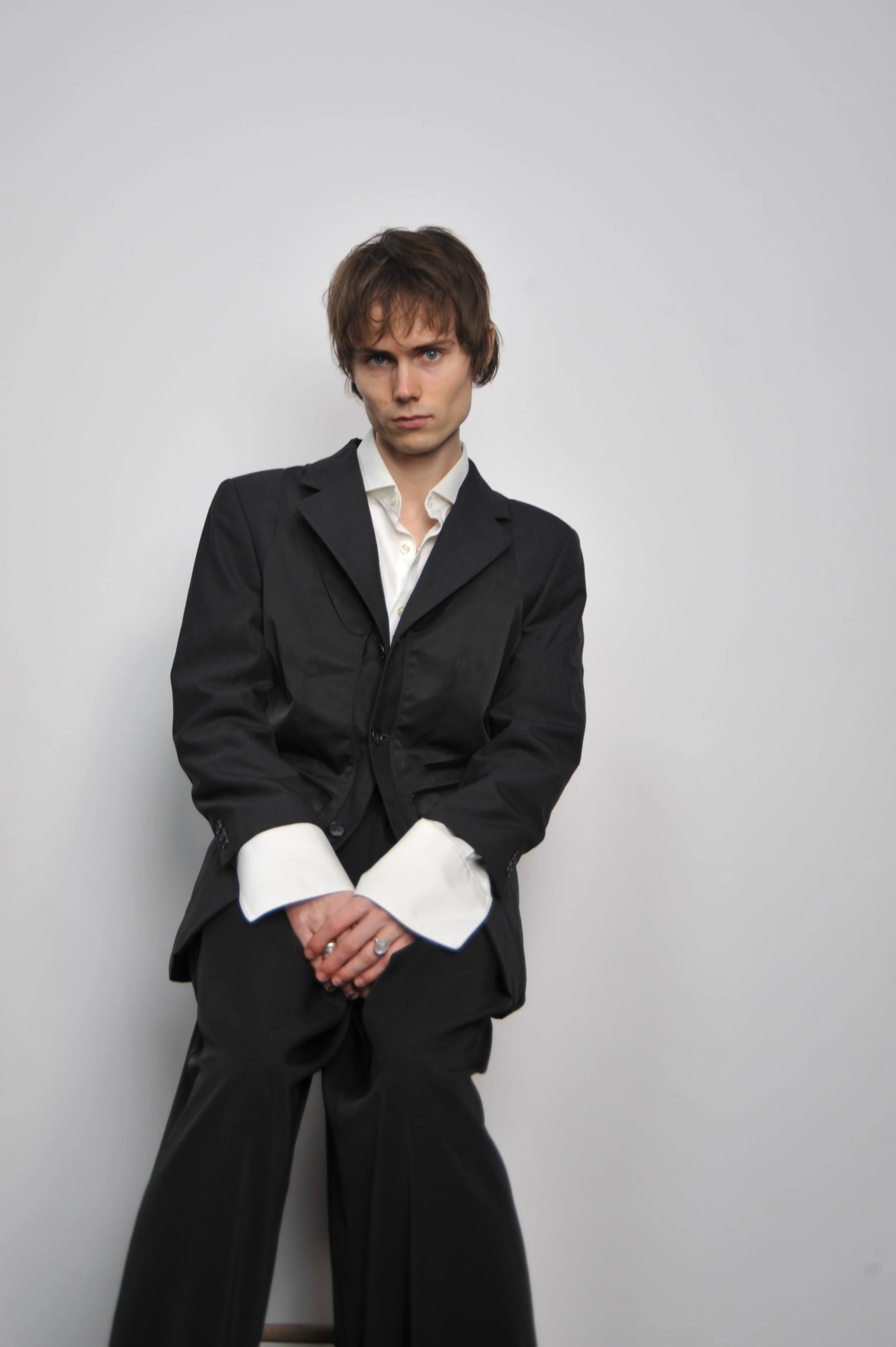 A young man with brown hair and blue eyes wearing a black suit with a white shirt, standing in front of a plain light grey background.