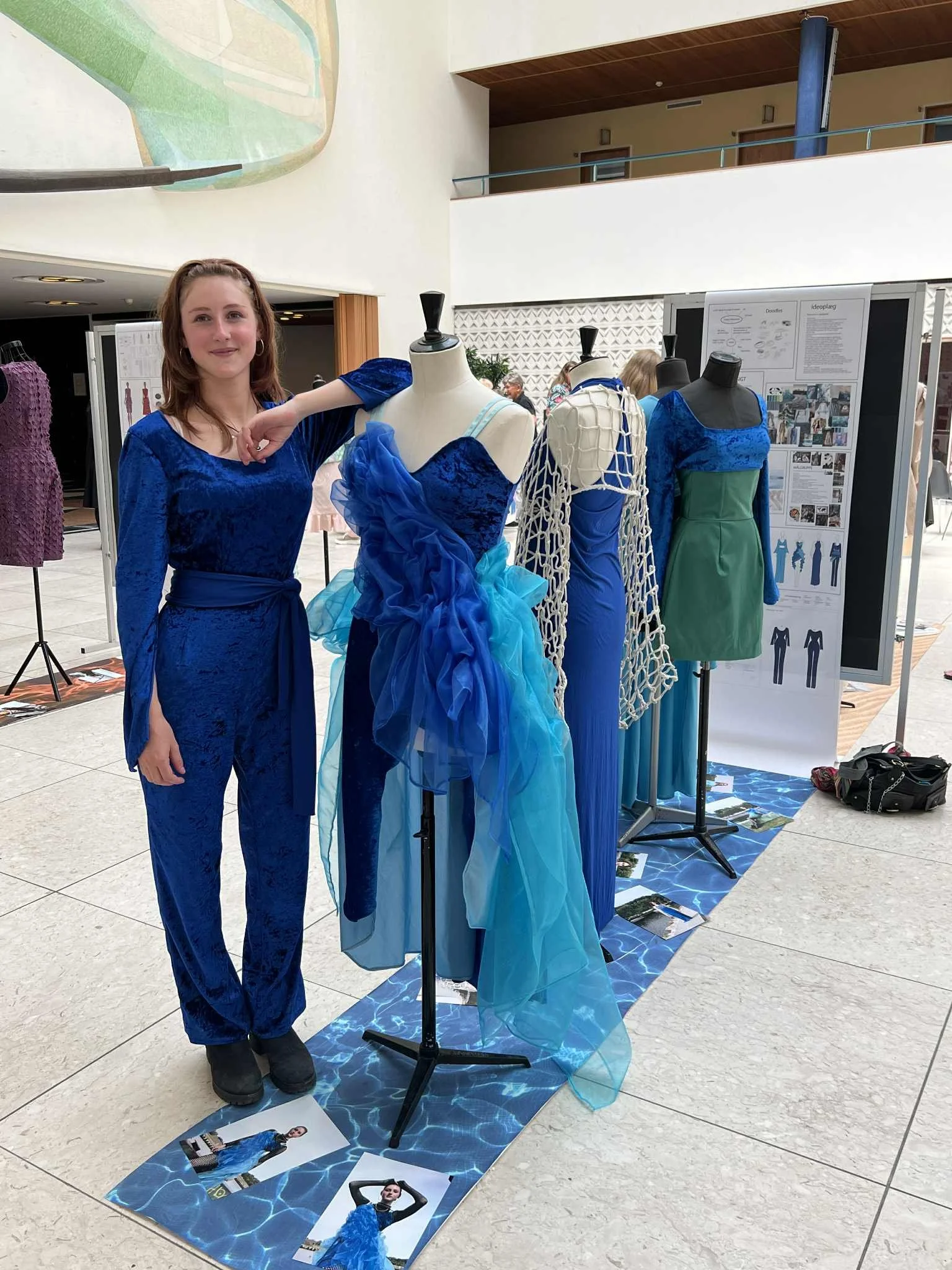 Young woman in a blue velvet jumpsuit standing next to dress mannequins displaying blue and green dresses at an indoor exhibition.