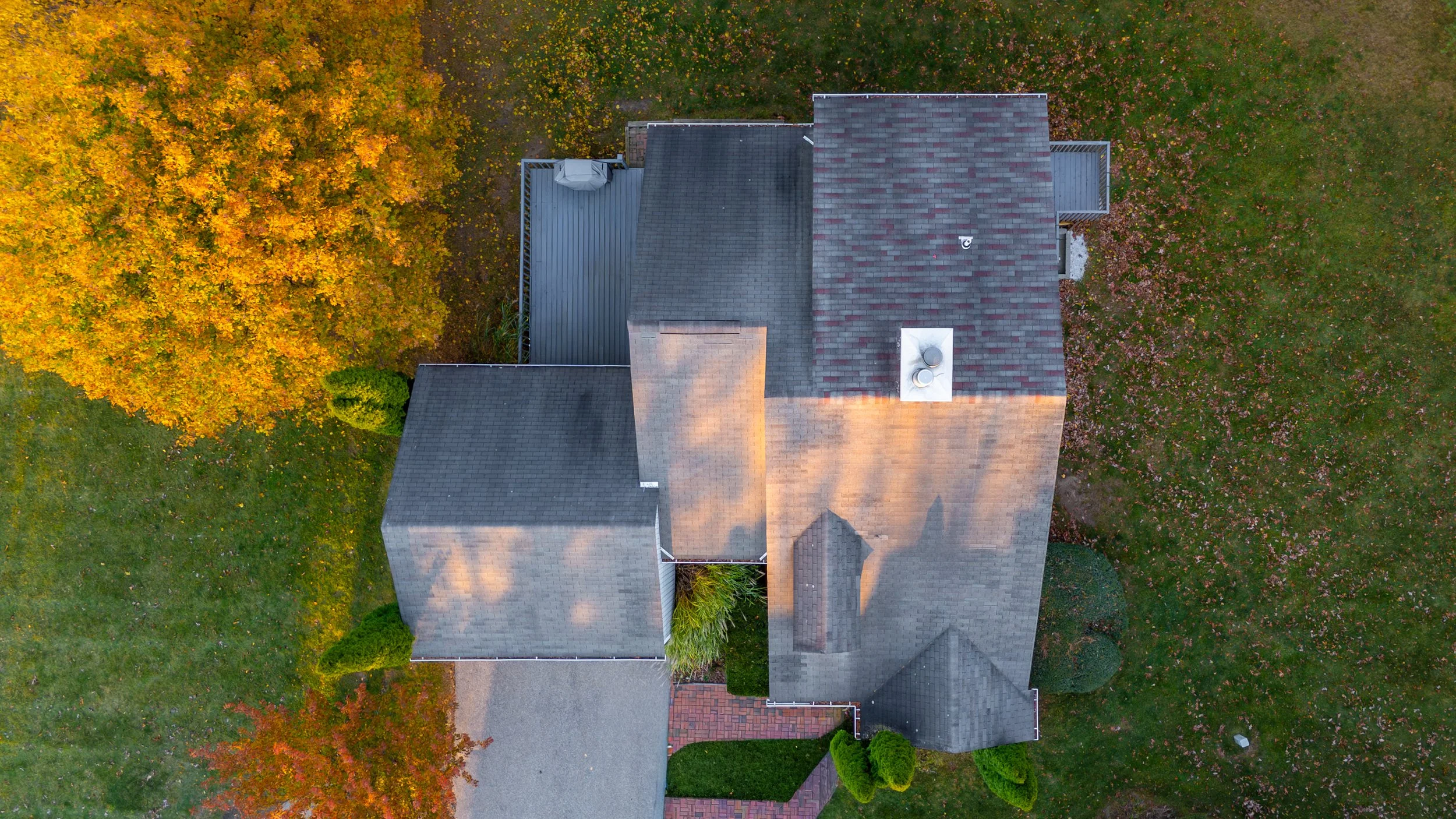 An aerial view of a house with a gray roof, surrounded by a lawn with autumn-colored trees and shrubs.
