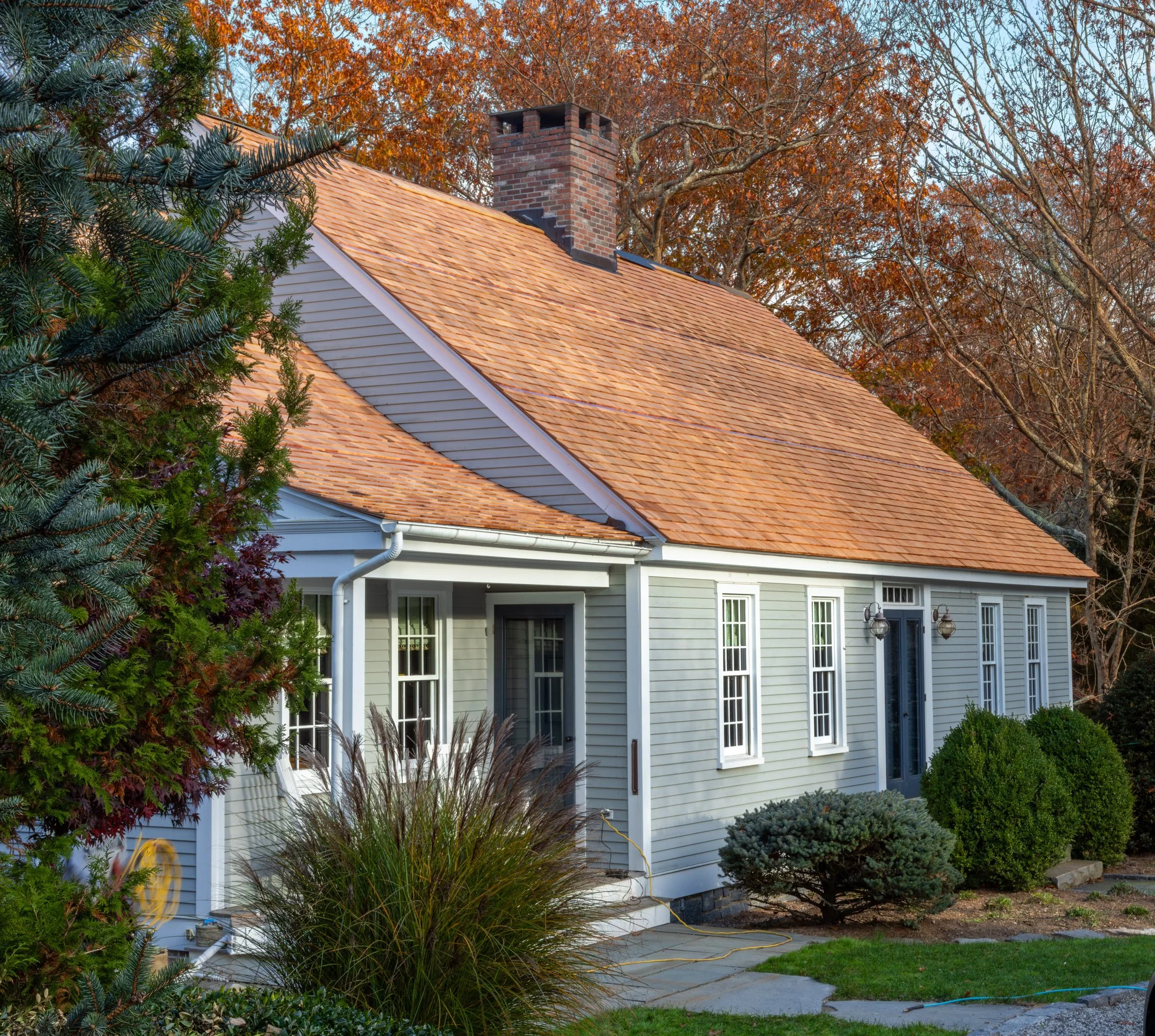 A house with a steeply pitched roof covered in orange tiles, gray siding, white trim, multiple tall narrow windows, and a brick chimney. There are trees and bushes in the yard, with autumn-colored leaves on the trees in the background.