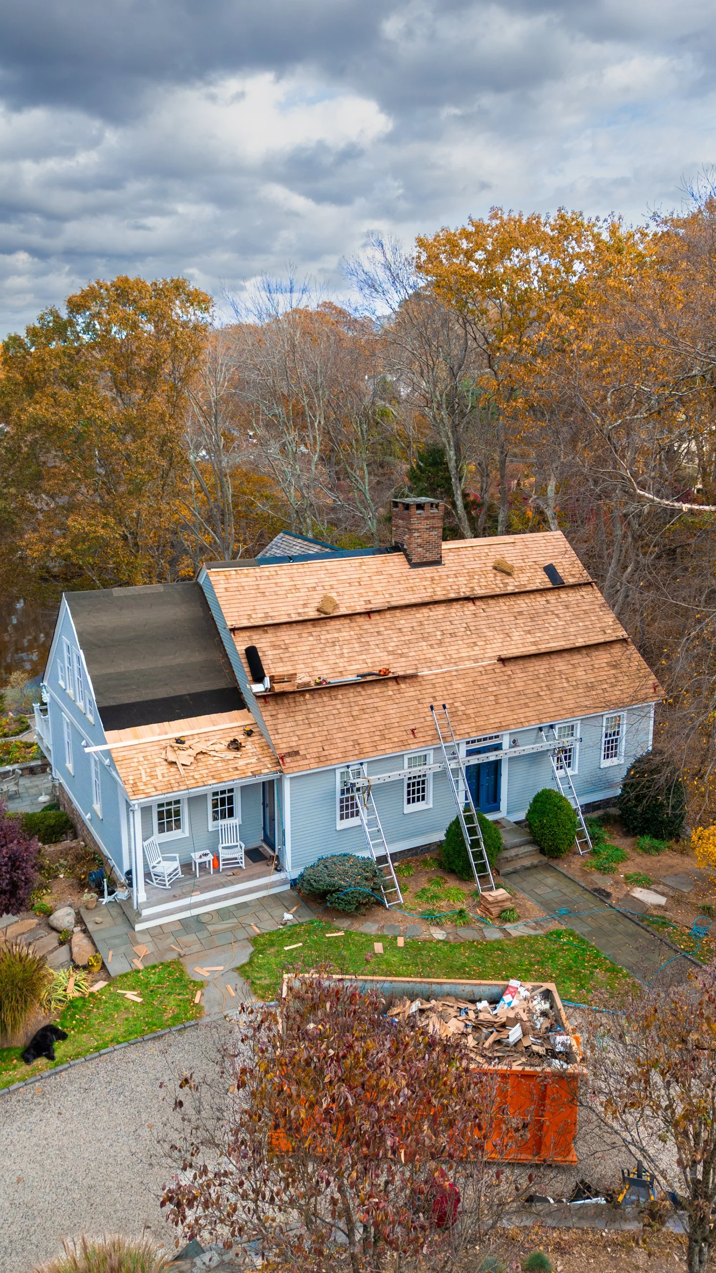 A house under renovation with a partially, newly installed shingle roof, ladders leaning against the second story, construction debris in a dumpster, and fall foliage surrounding the property.