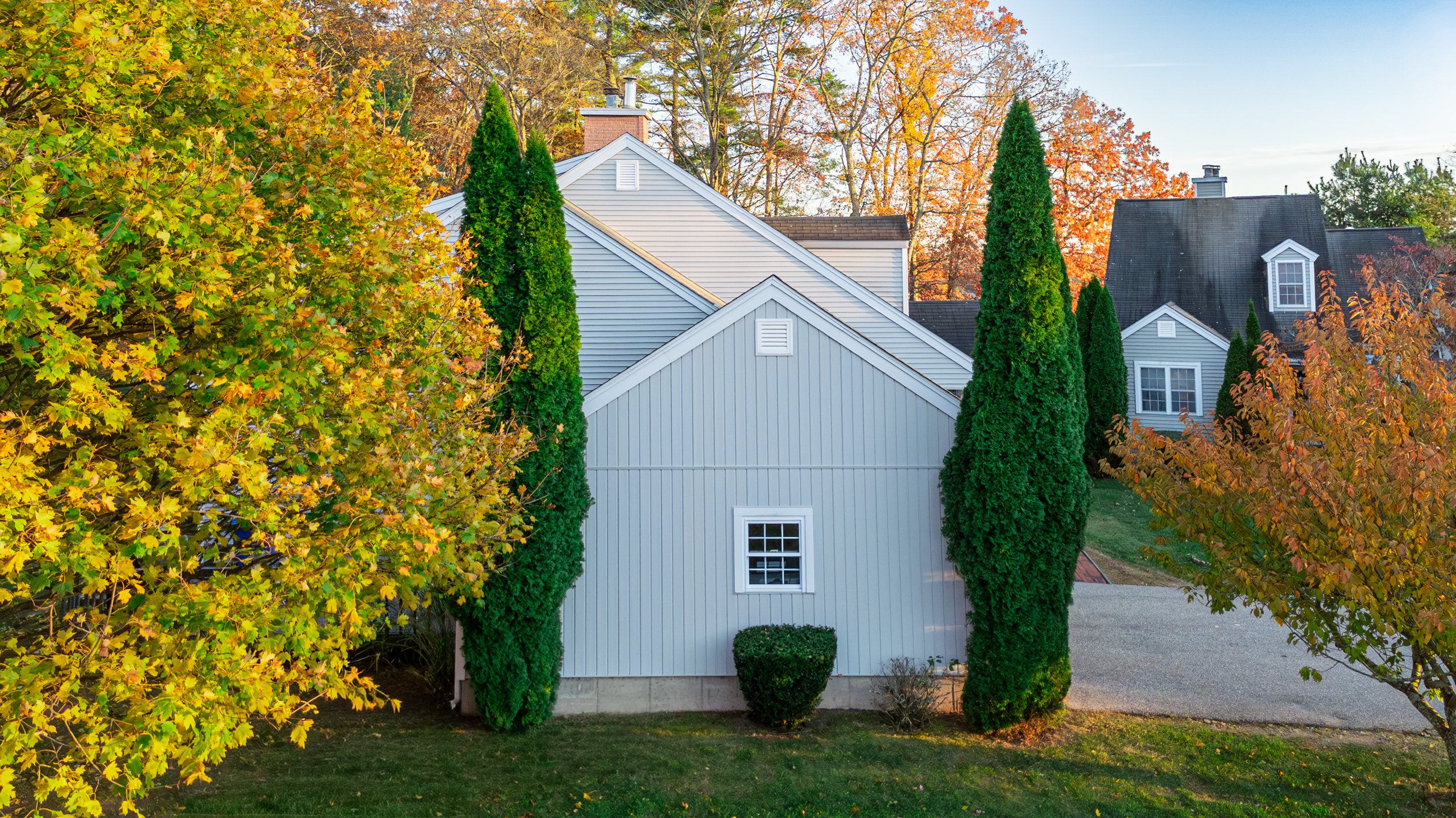 A white house with vertical siding is surrounded by trees with autumn leaves and tall green conifers, with a clear blue sky in the background.