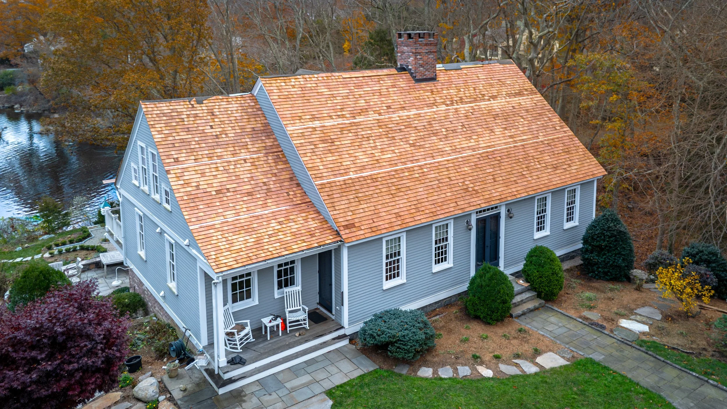 Aerial view of a gray house with a red tile roof, surrounded by a landscaped yard with trees and shrubs, adjacent to a water body in the background.