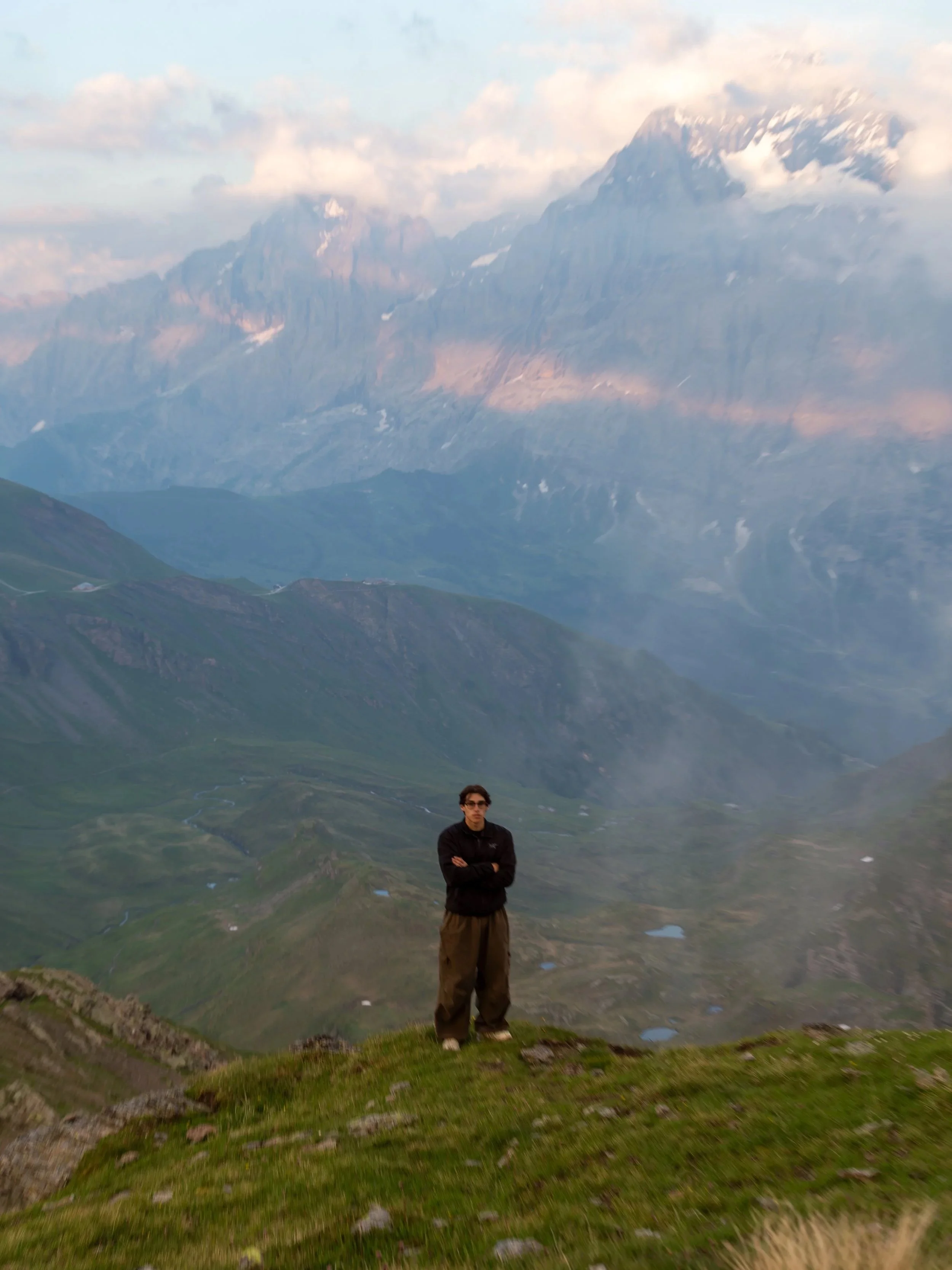 A person standing on a grassy hillside in a mountainous landscape with large snow-capped mountains and clouds in the background.