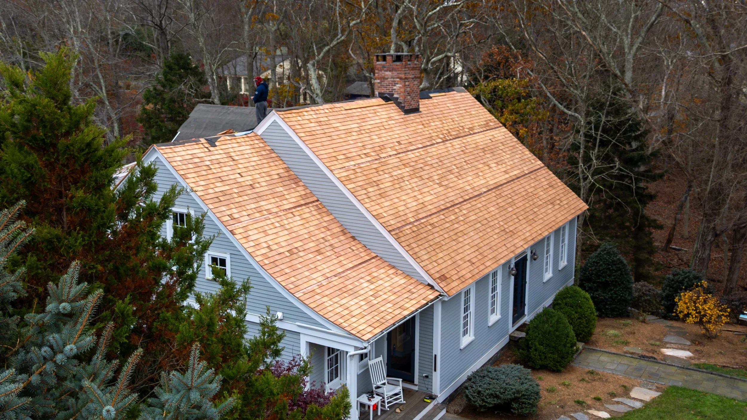 An aerial view of a house with new wooden shingle roofing being installed, surrounded by trees and shrubs.