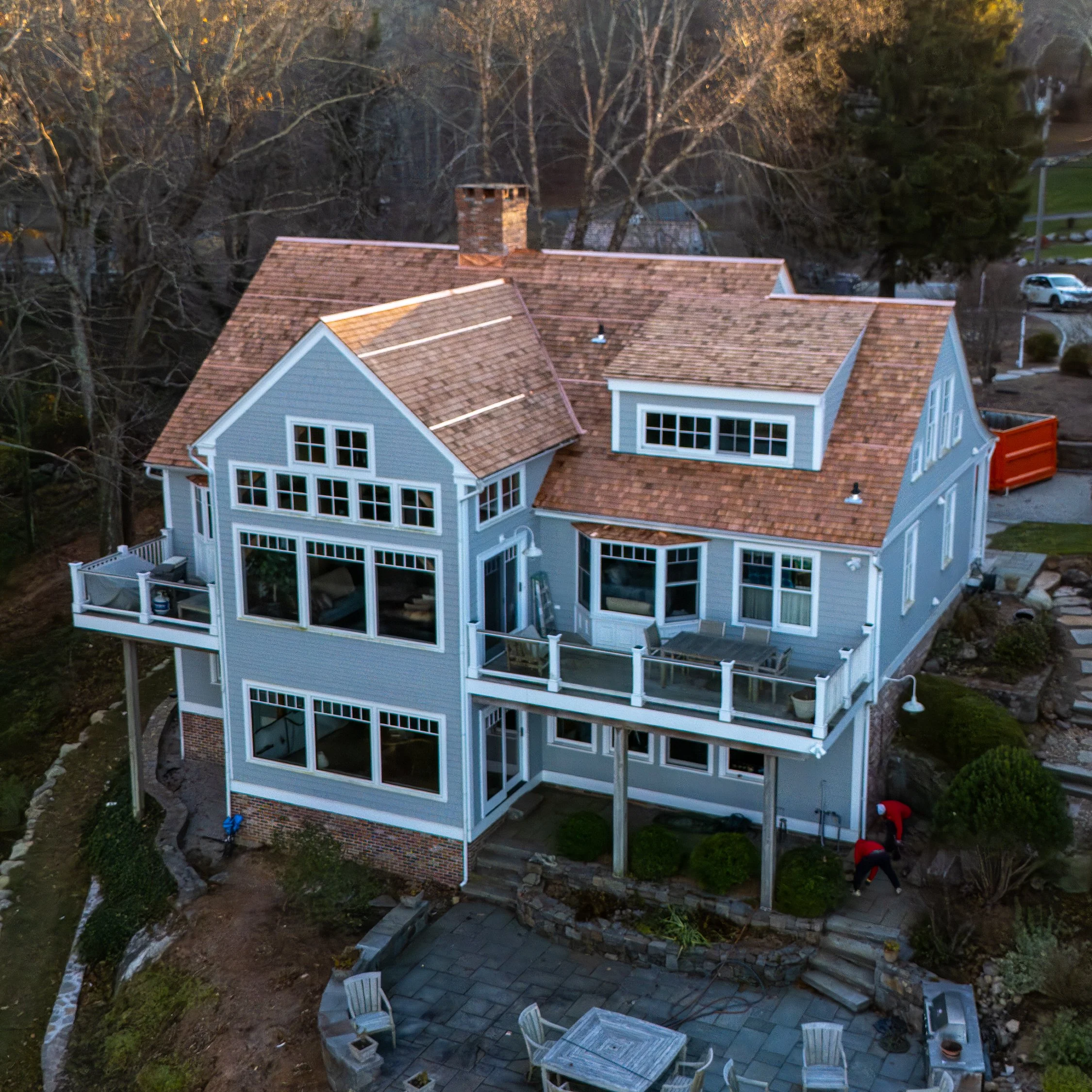 An aerial view of a blue multi-story house with a brown tiled roof, multiple large windows, and a spacious deck with outdoor furniture. There are two people working near the garden, and the yard features a patio area with chairs and a table.