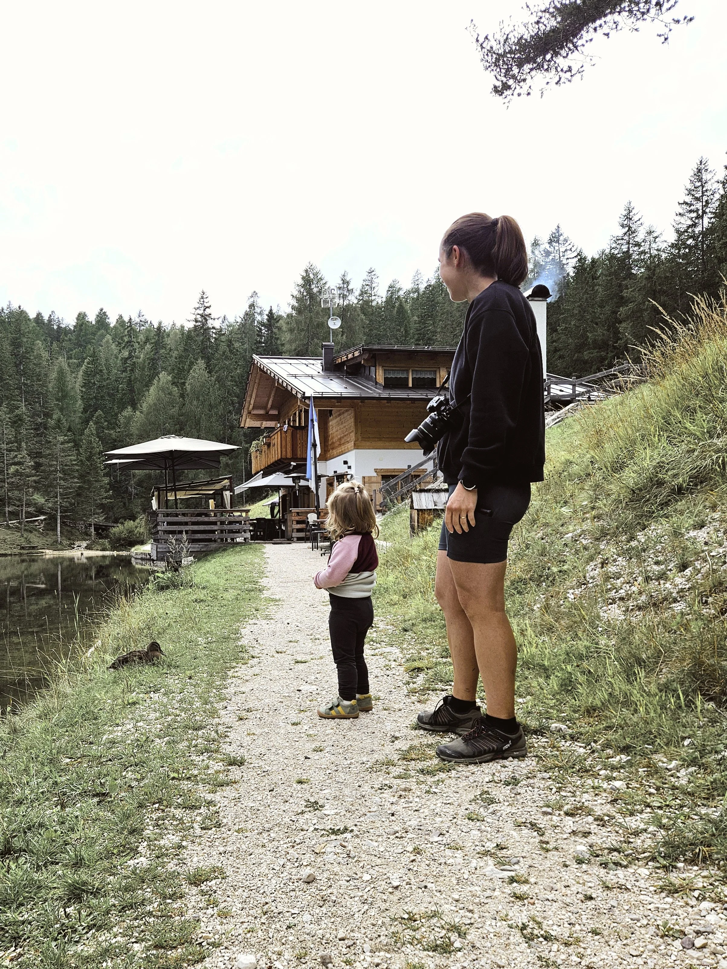 Žena s dítětem stojící na pěšině na břehu jezera, s pohledy směřujícími k roubené chatě uprostřed přírody.