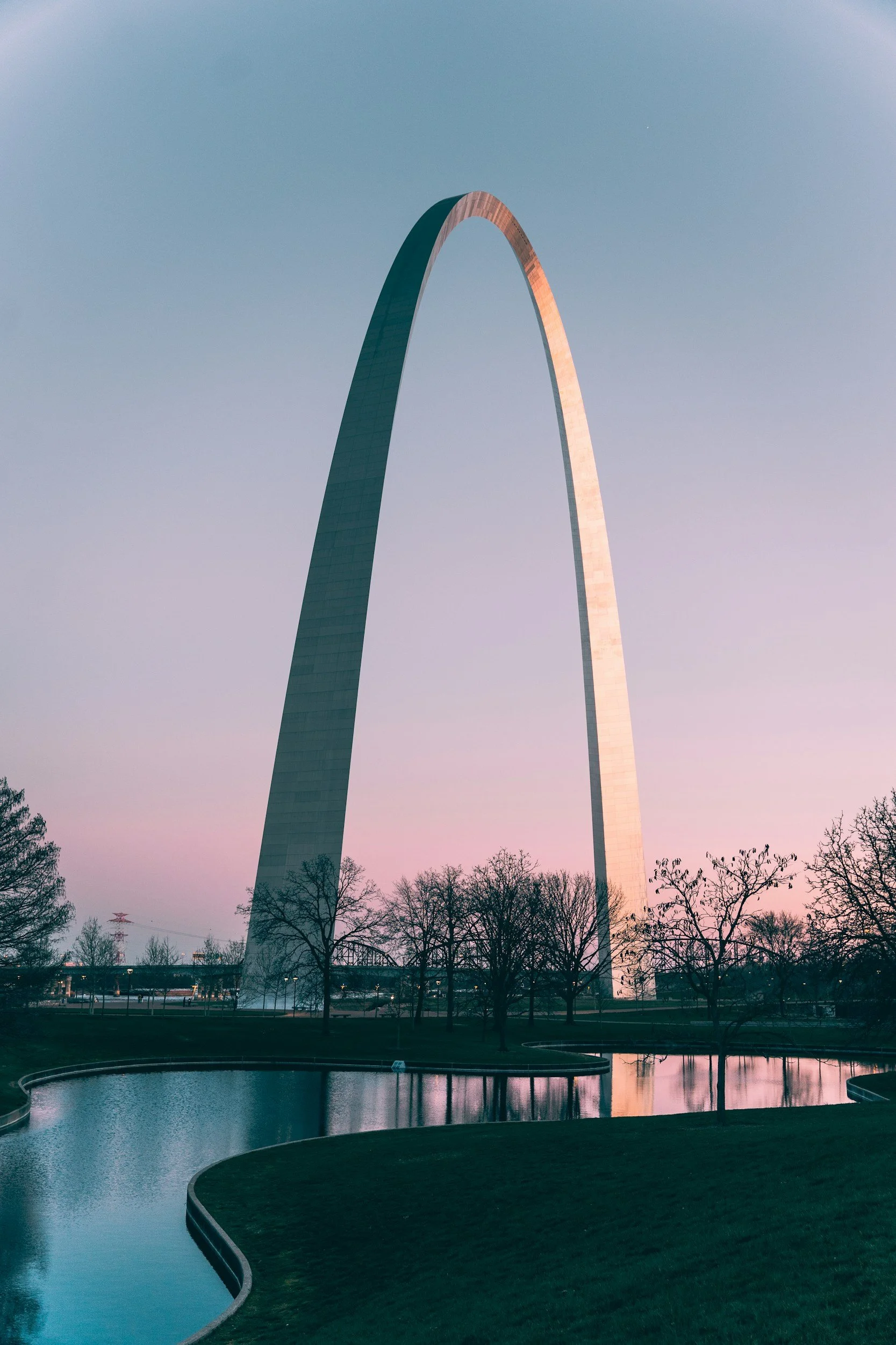 The Gateway Arch in St. Louis, Missouri, during sunset, with pink and purple hues in the sky and trees reflected in the water in the foreground.