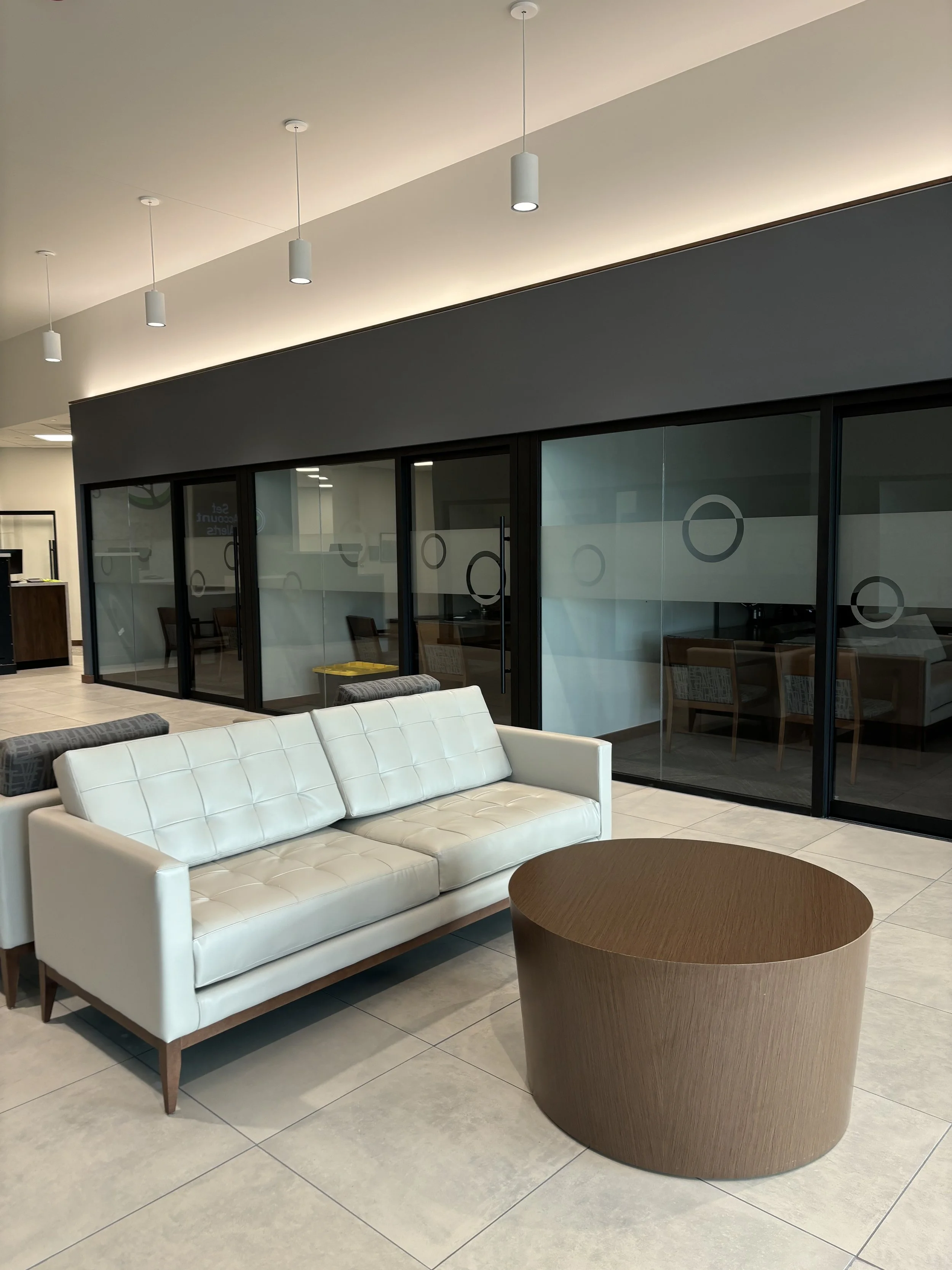 Modern  office lobby with a white leather sofa, round wooden coffee table, and glass-walled meeting rooms in the background.